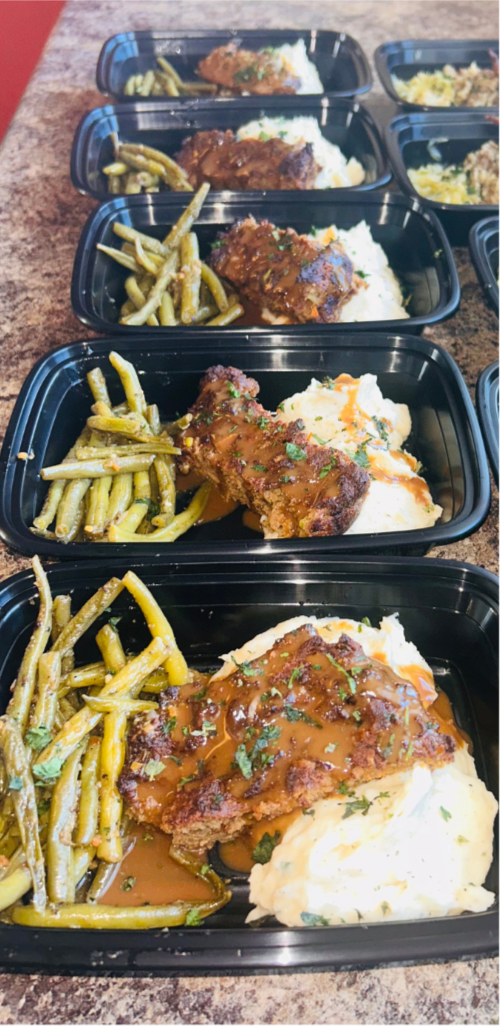 Black meal prep containers with mashed potatoes, fried chicken with gravy, and green beans, arranged on a brown countertop.