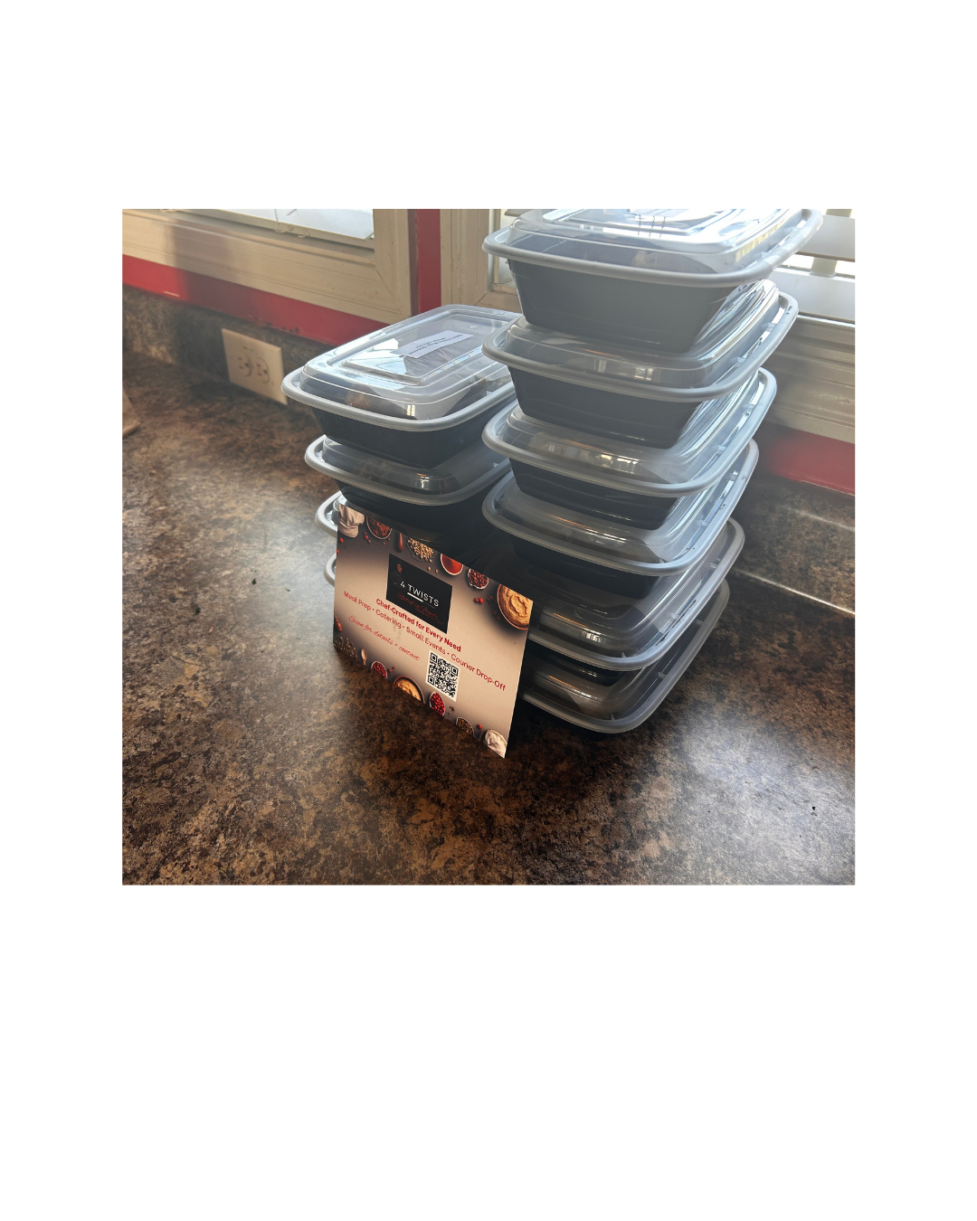 Stacked clear food containers with black bases on a countertop near a window, with a promotional sign for a catering service.