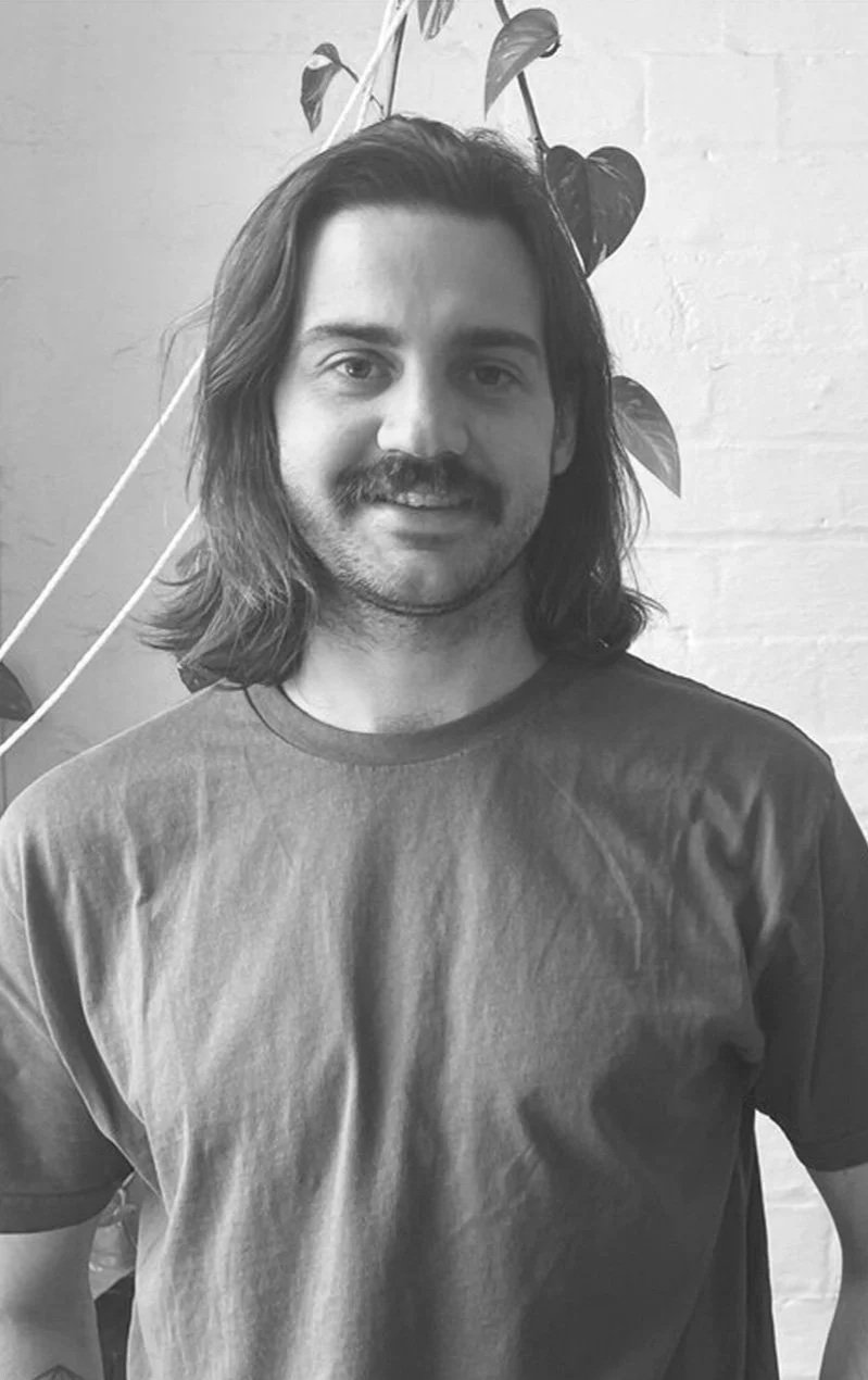A man with long hair and a mustache standing in front of a white brick wall with a plant behind him.