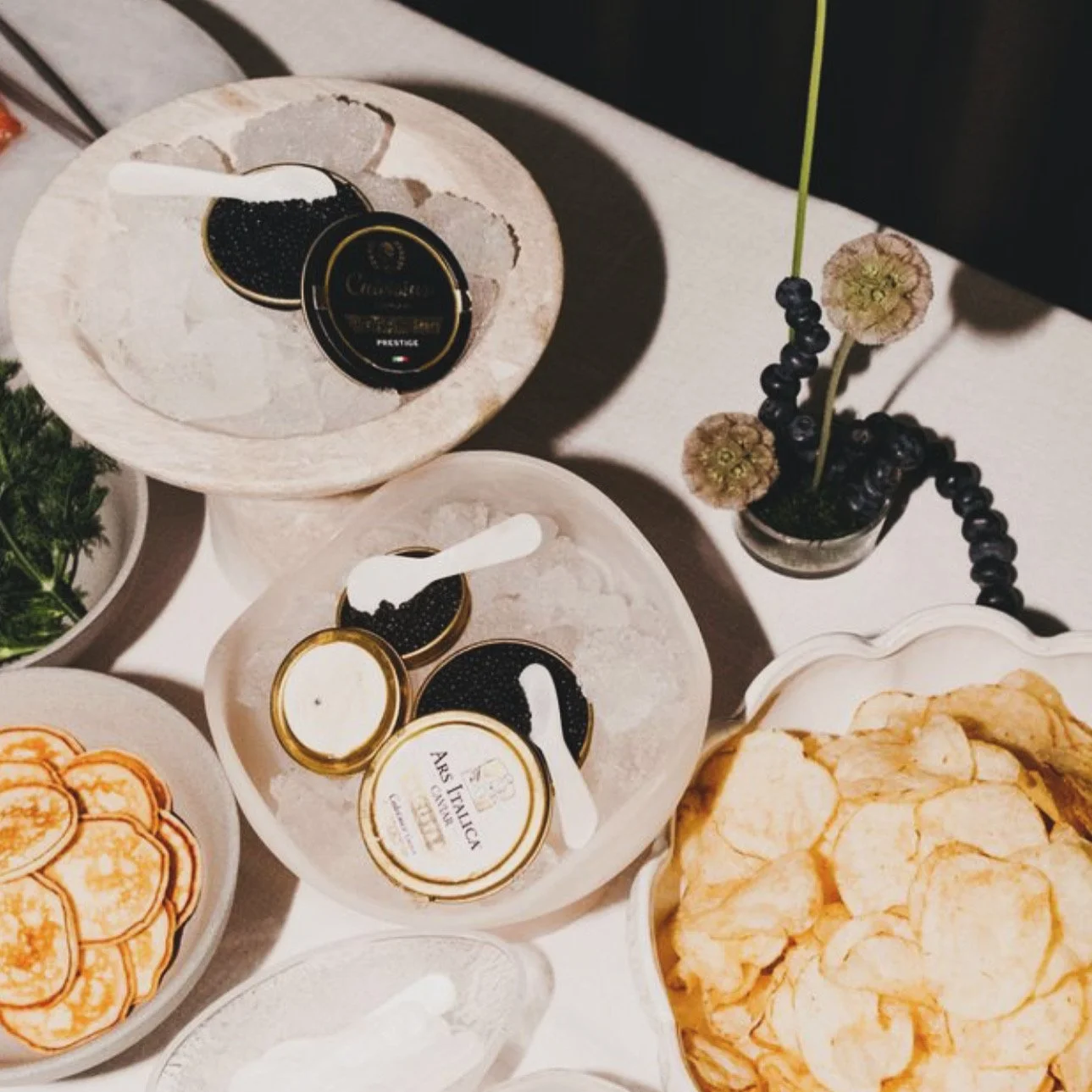 A table with bowls of potato chips, caviar containers with spoons, and a small vase with black currants and dried flowers.