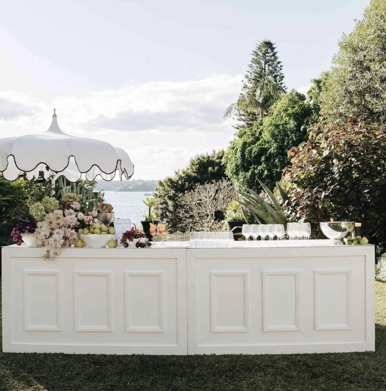 Mobile bar setup with glassware and floral decorations under a white umbrella, overlooking a body of water and lush trees.