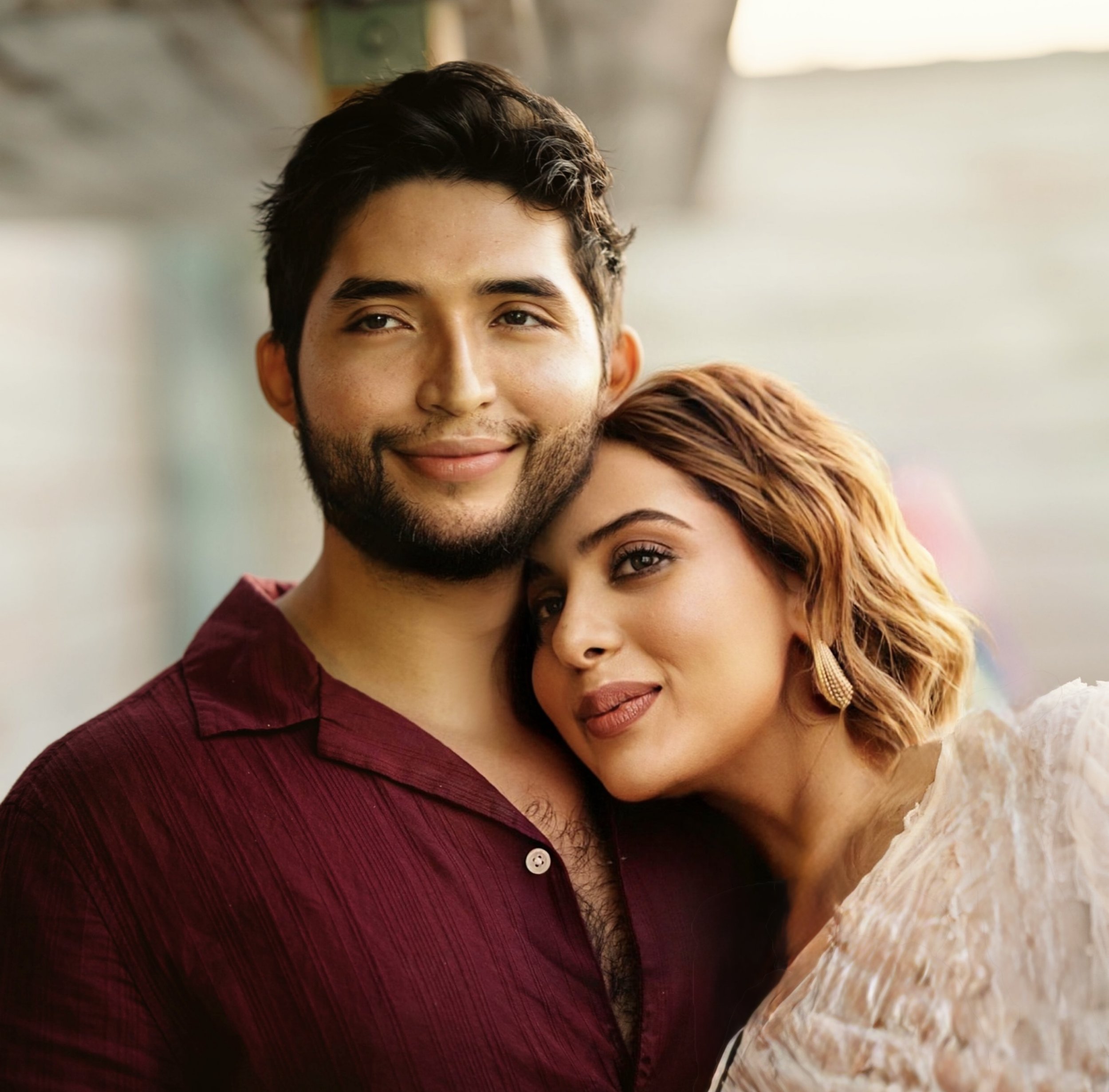 A happy couple embraces, the man with dark hair and a beard wearing a maroon shirt, and the woman with reddish-brown hair and gold earrings wearing a white textured top, standing close together and smiling.