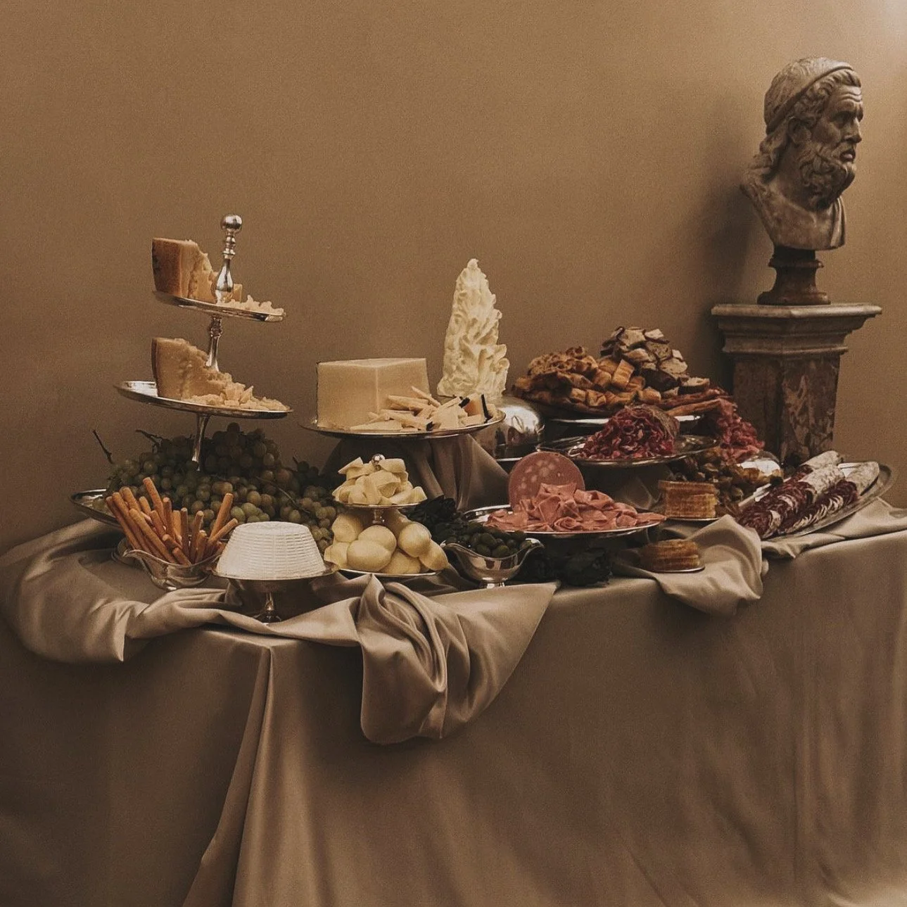 A lavish table spread featuring cheeses, grapes, meats, breadsticks, and various charcuterie items in a dimly lit room with classical sculptures.