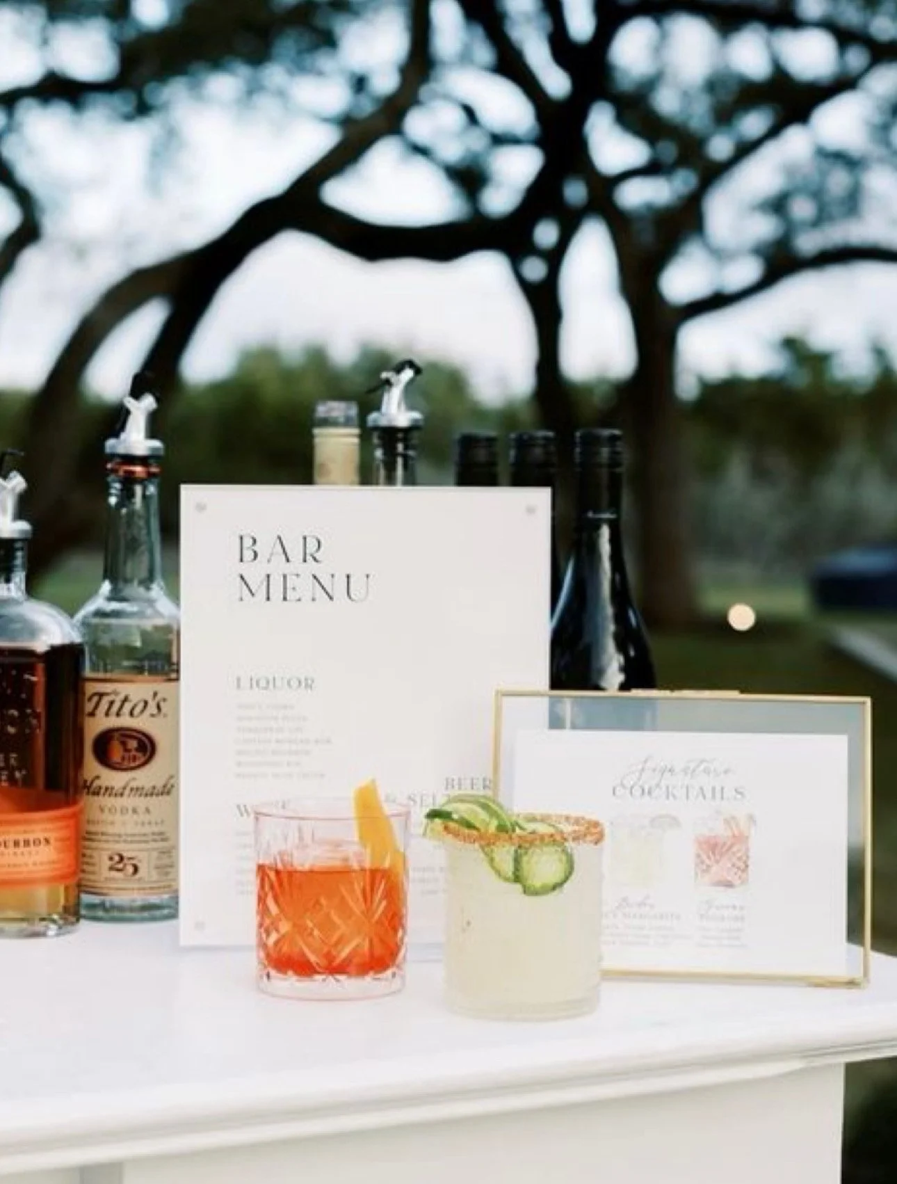 Bar setup with bottles of liquor, drink menu, and two cocktails on a table outdoors, with trees in the background.