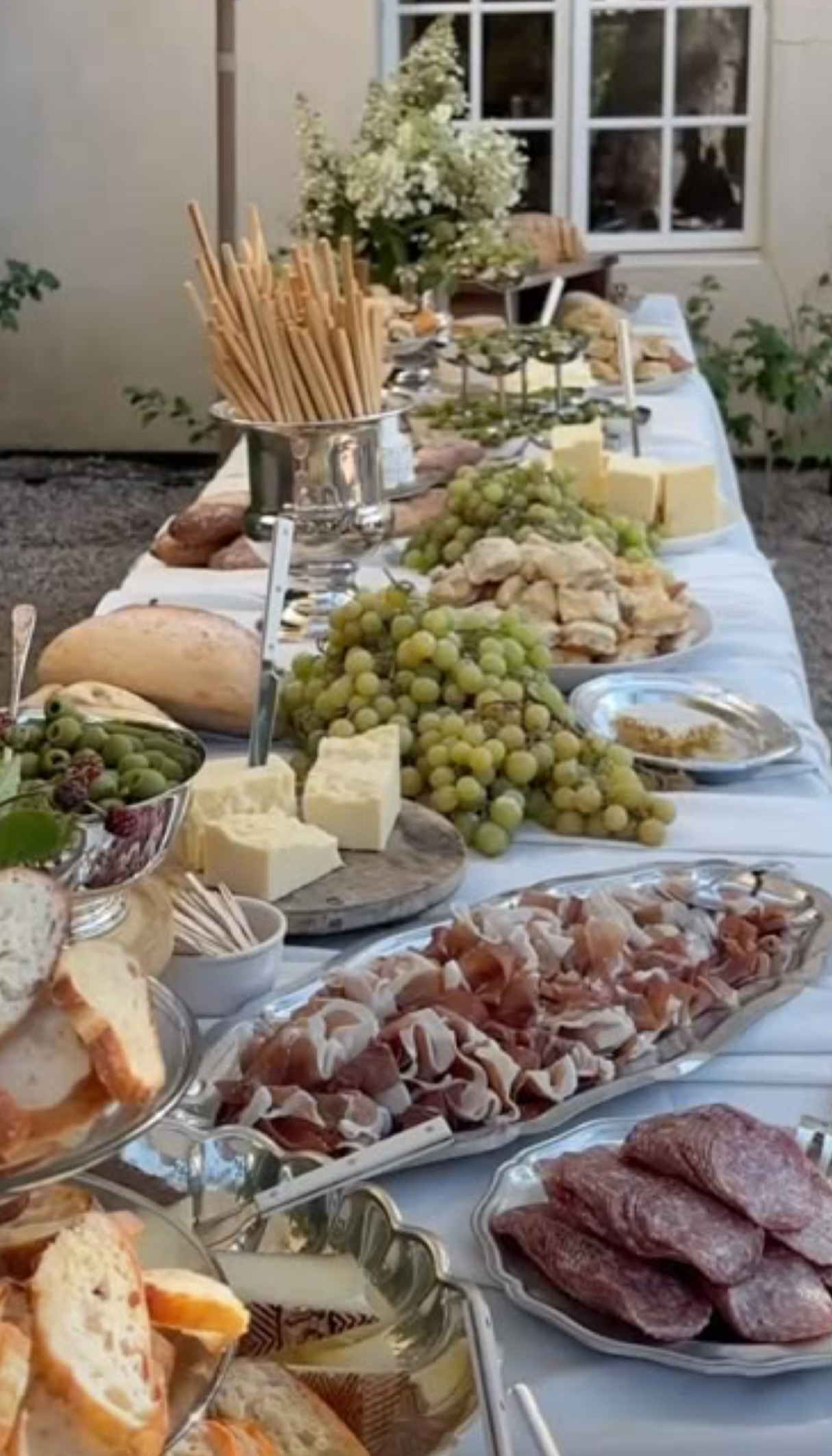 Table set with various cheeses, grapes, meats, bread, and crackers, outdoors near a house with a window.