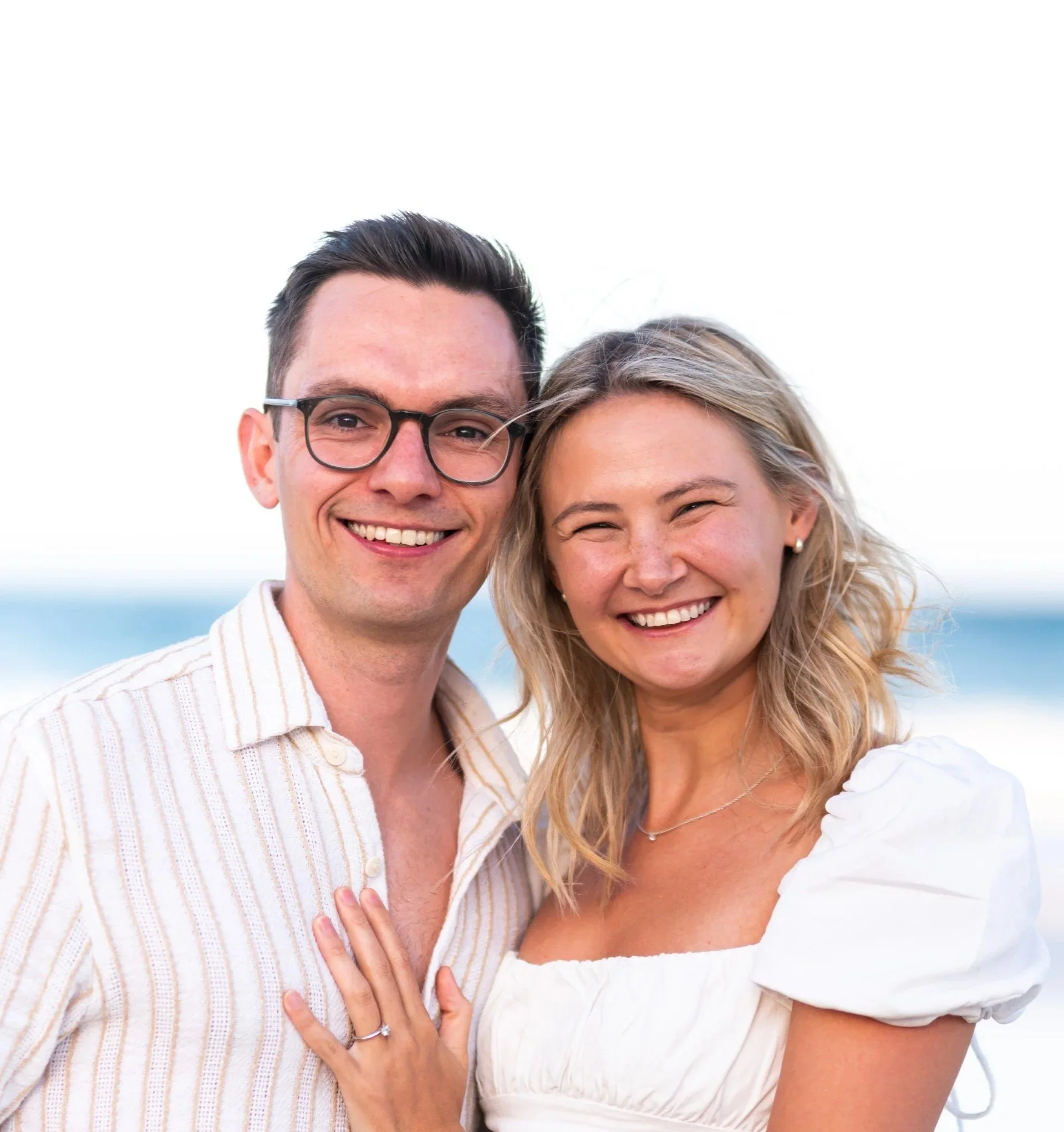 A smiling couple at the beach, with the ocean in the background, standing close together and showing off an engagement ring.