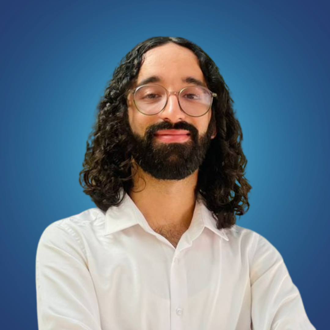 A man with long curly hair, glasses, and a beard, wearing a white shirt, standing against a blue background.