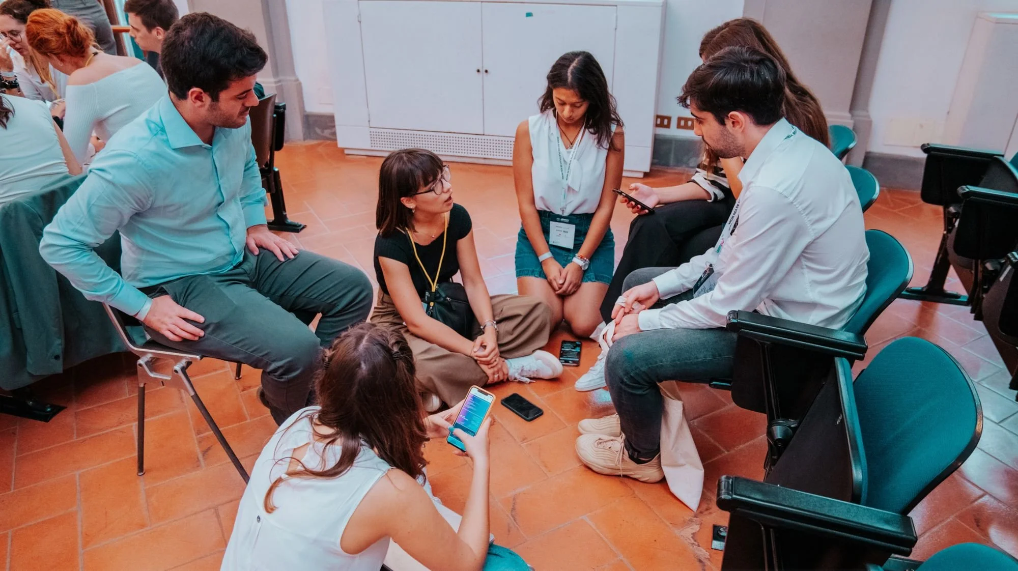 A group of young people sitting on the floor in a circle, engaging in conversation, with some holding their phones.