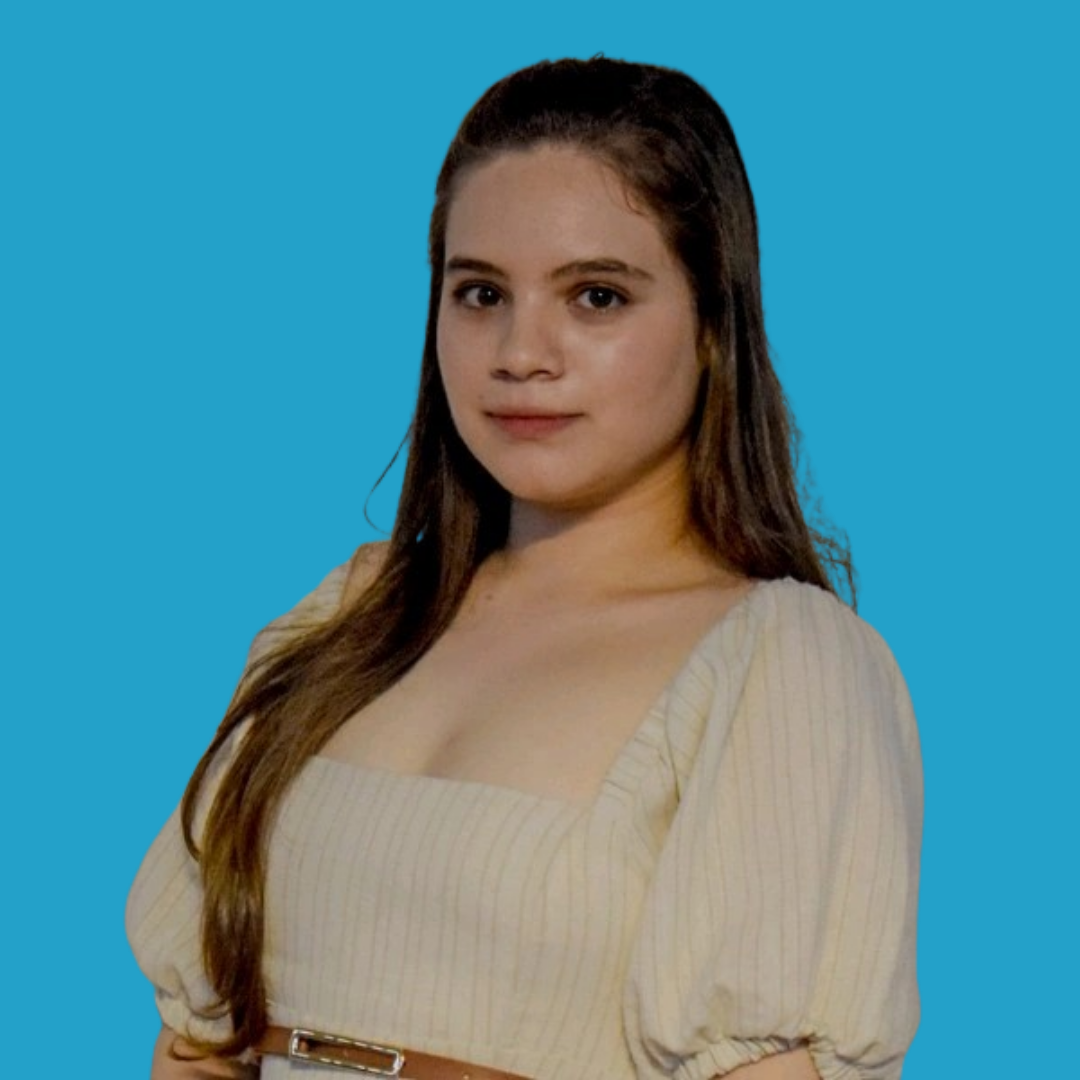 Portrait of a young woman with long brown hair wearing a beige top, standing against a blue background.