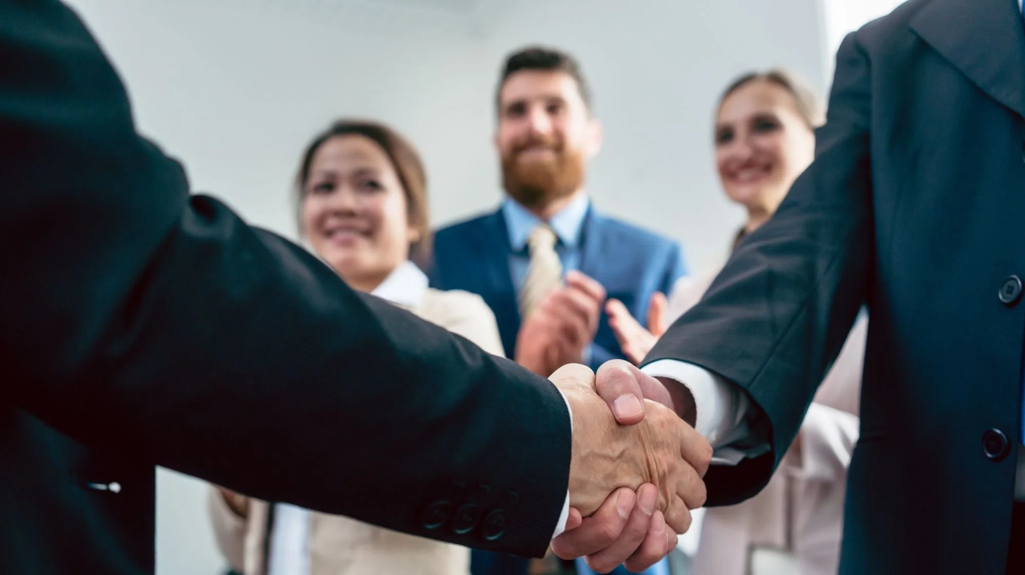 Close-up of a handshake between two businesspeople, with three colleagues clapping and smiling in the background in a professional setting.