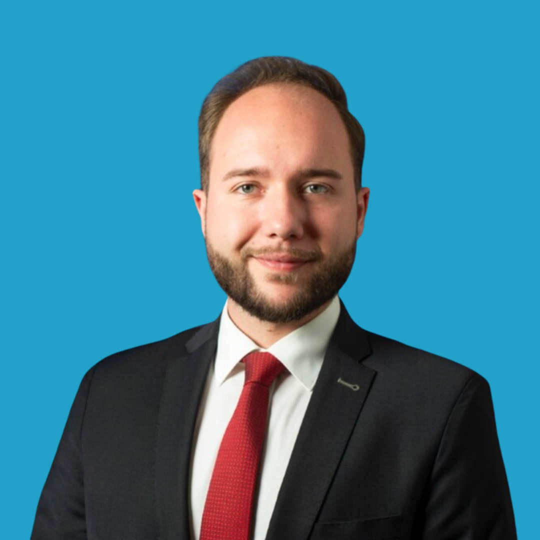 Professional headshot of a man with light skin, brown hair, and a beard, wearing a black suit, white shirt, and red tie, against a solid blue background.