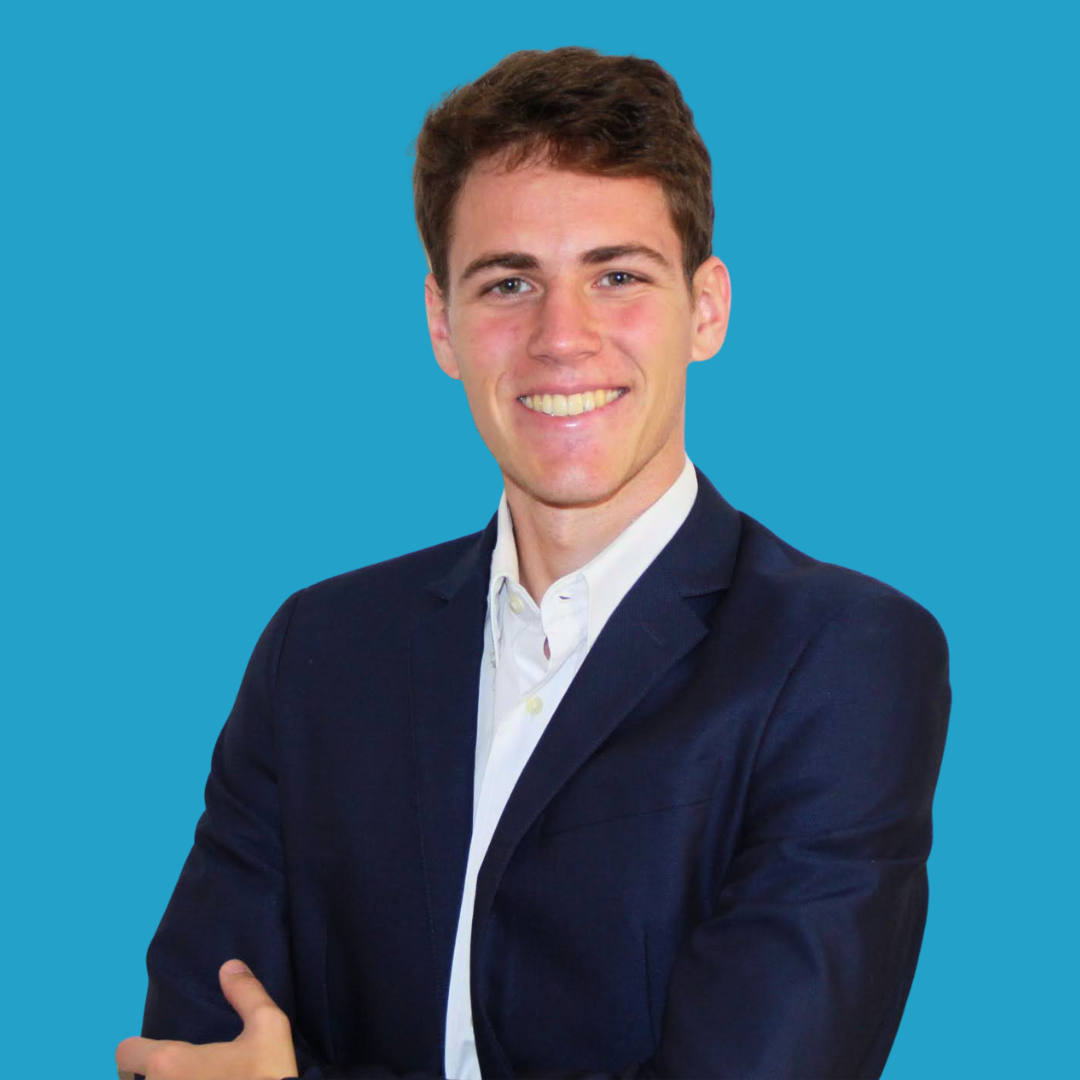 A young man with short brown hair, wearing a white shirt and navy blazer, smiling with arms crossed, against a solid blue background.
