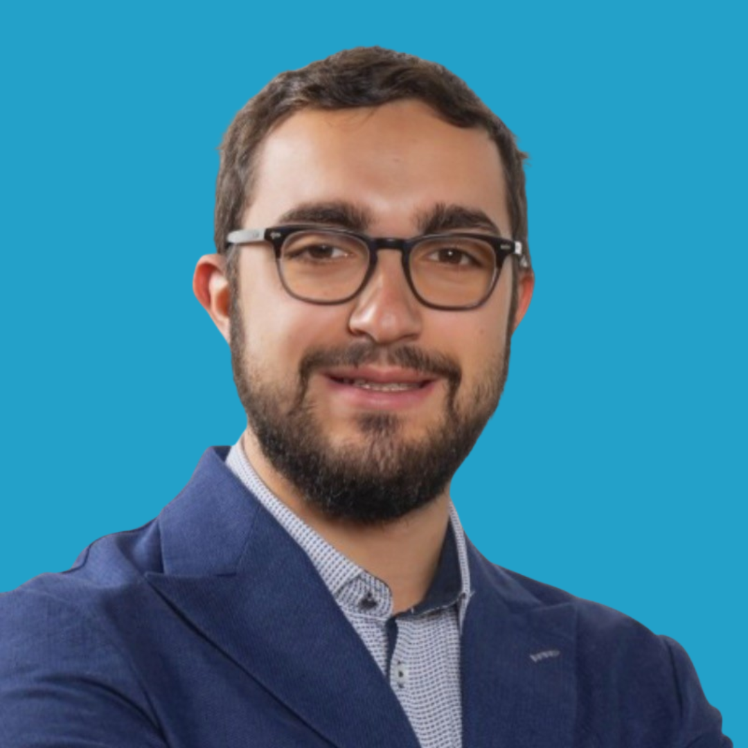 A young man with glasses, a beard, and short hair, wearing a navy blazer and a button-up shirt, smiling against a solid blue background.