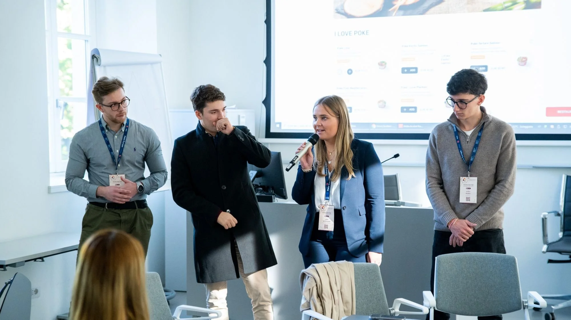 Four young professionals at a conference or workshop, standing in front of a large screen displaying a website or presentation, with some audience members seated in the foreground.