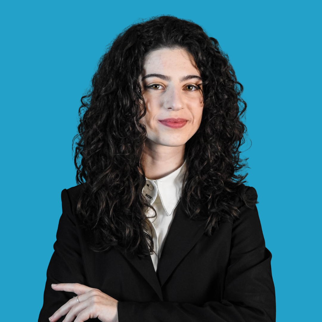 A woman with curly dark hair smiling, wearing a black blazer and a white collared shirt, against a blue background.