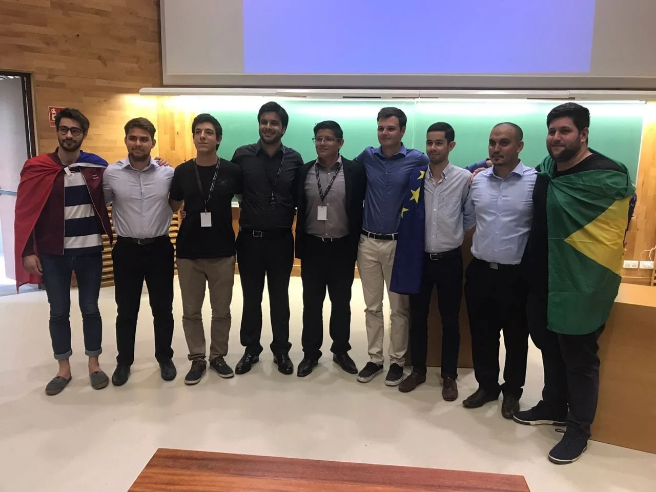 Group of ten men standing together in a classroom or conference room, some with flags draped over their shoulders, smiling and posing for a photo.