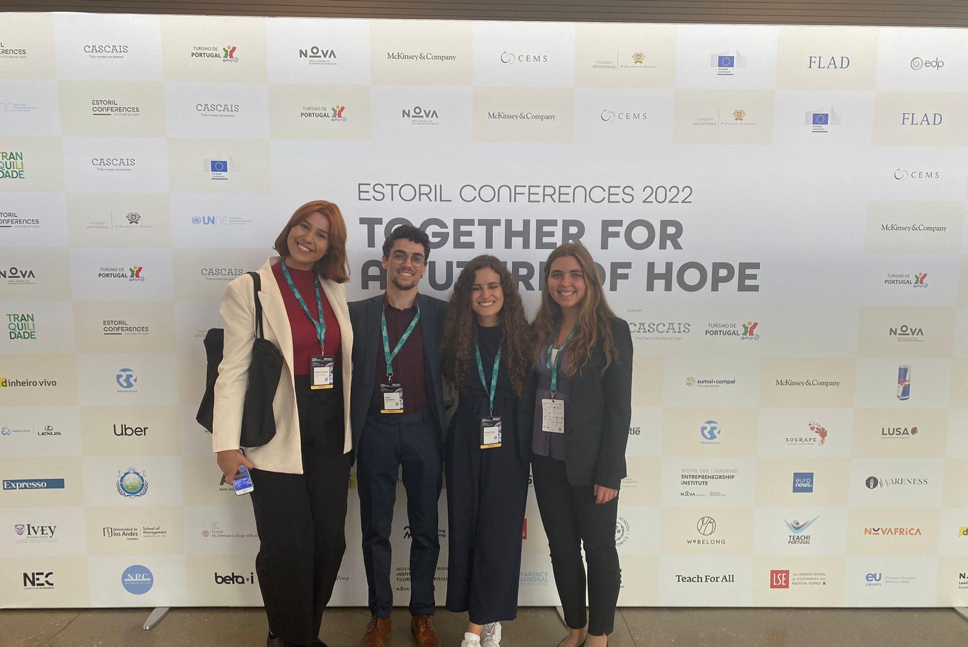 Four smiling young adults standing in front of a conference backdrop at the Estoril Conferences 2022, dressed in business casual attire with conference badges.