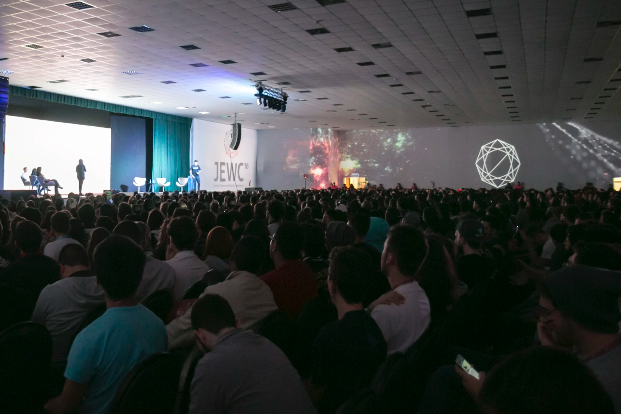 Large crowd attending a conference with a stage showing the event name "JEWC 16" and colorful digital graphics on the screen.