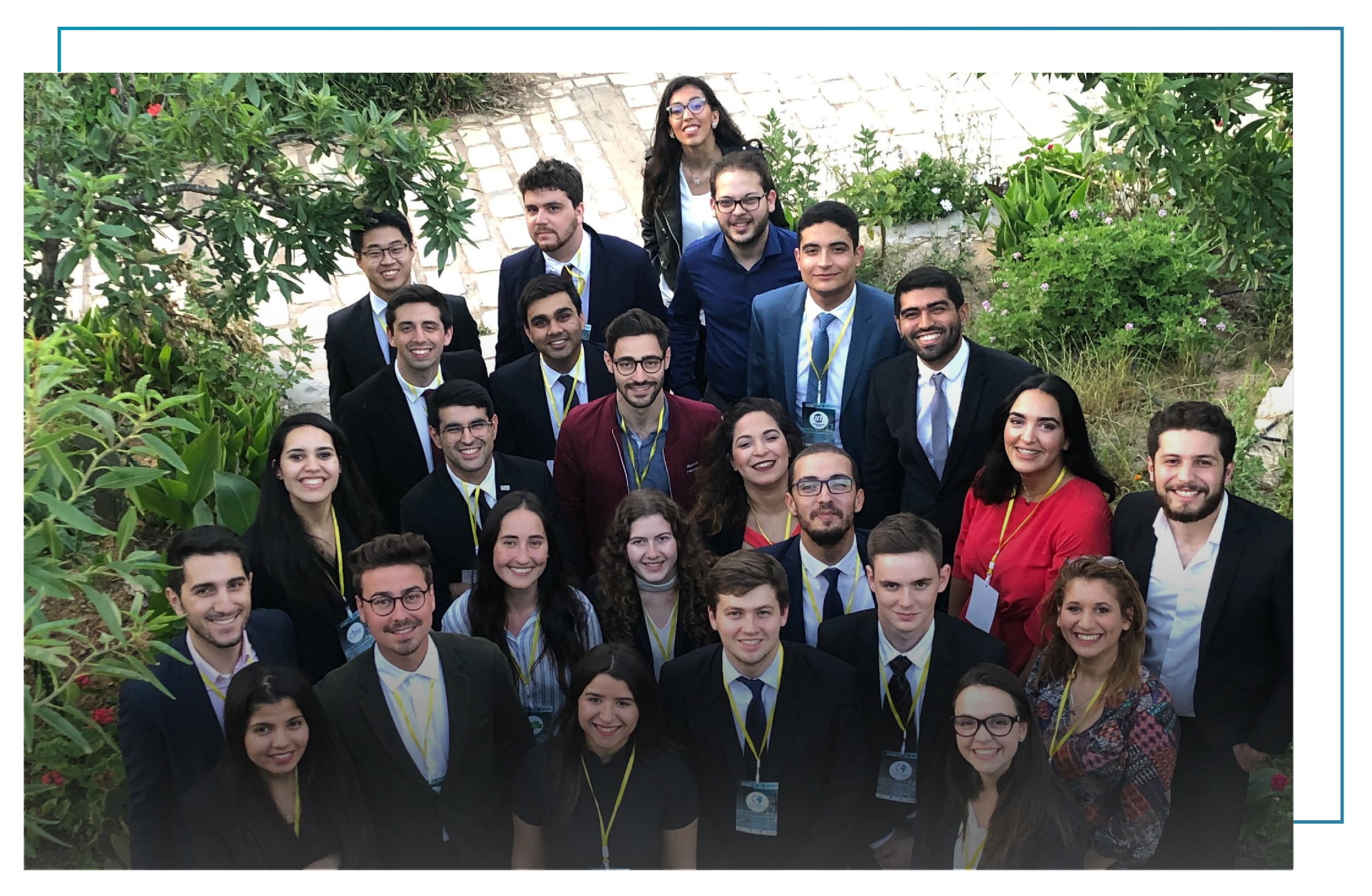 Group of young professionals at a corporate event, outdoors with plants and a stone pathway, smiling for the camera.