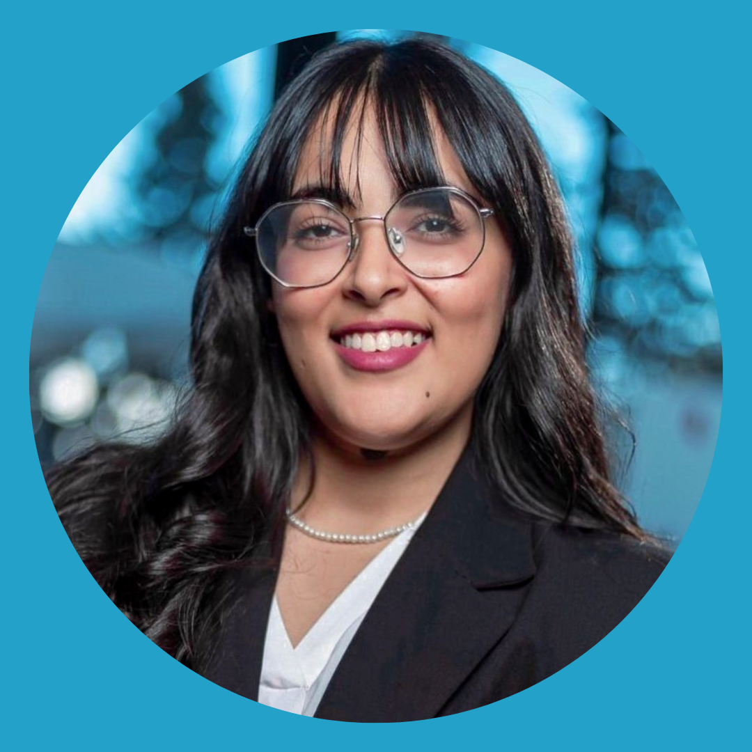 A woman with long dark hair, glasses, and a pearl necklace, smiling outdoors with a blurred blue background.