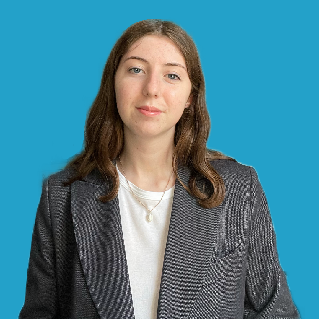 A young woman with shoulder-length brown hair, wearing a dark blazer over a white top and a pearl necklace, standing against a solid blue background.