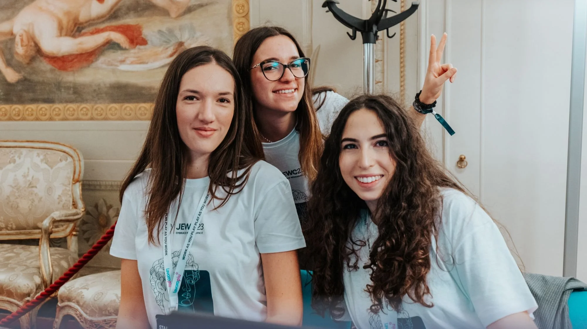 Three young women smiling and posing together indoors, with one making a peace sign, in a setting with ornate furniture and a painting on the wall.