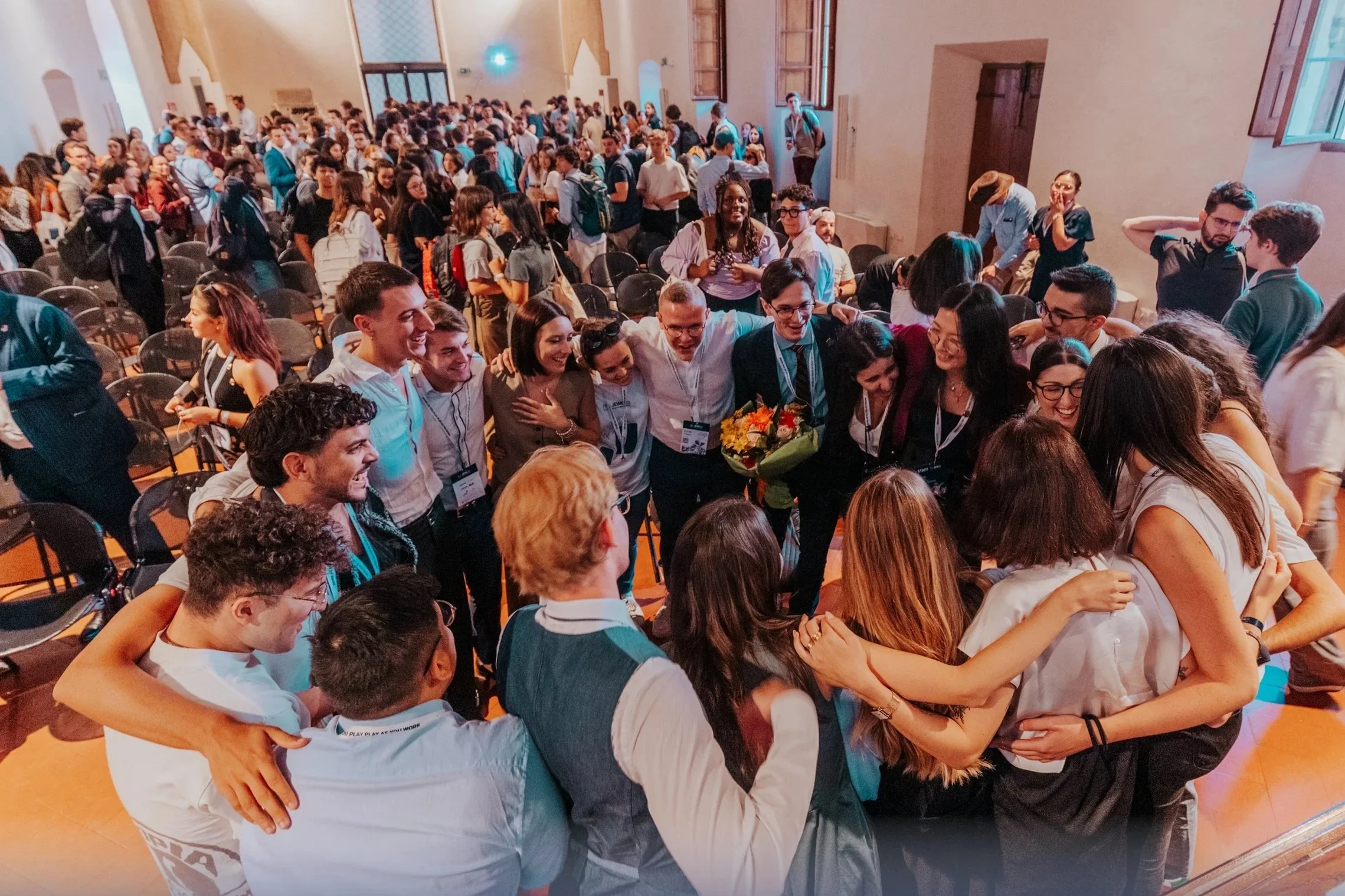 A group of people gathered indoors, hugging and celebrating. In the background, a large crowd is standing and sitting in a room with high windows and wooden accents.