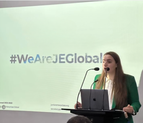 A woman speaking at a podium during a presentation, with a screen behind her displaying the hashtag '#WeAreJEG’. She has long brown hair and is wearing a green blazer.