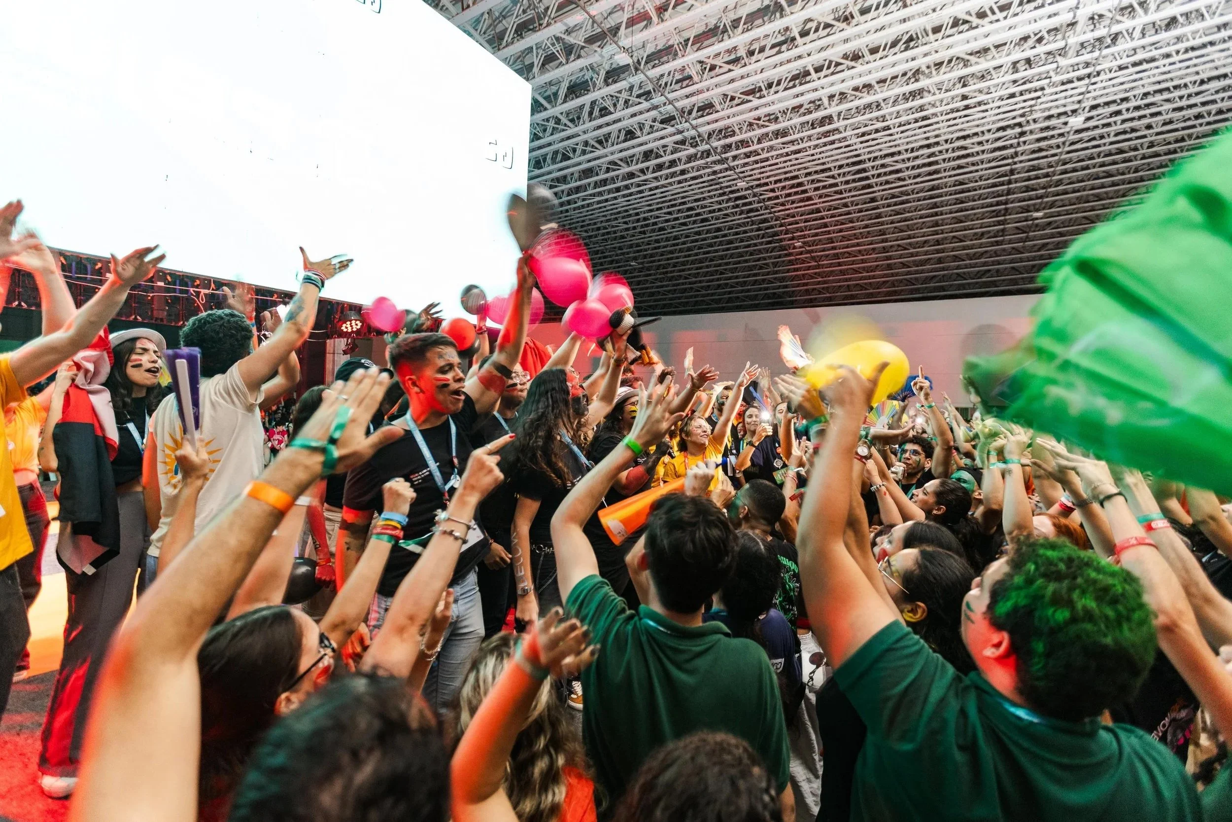 Crowd of people celebrating at an indoor event with colorful balloons, masks, and face paint, under a large ceiling structure.