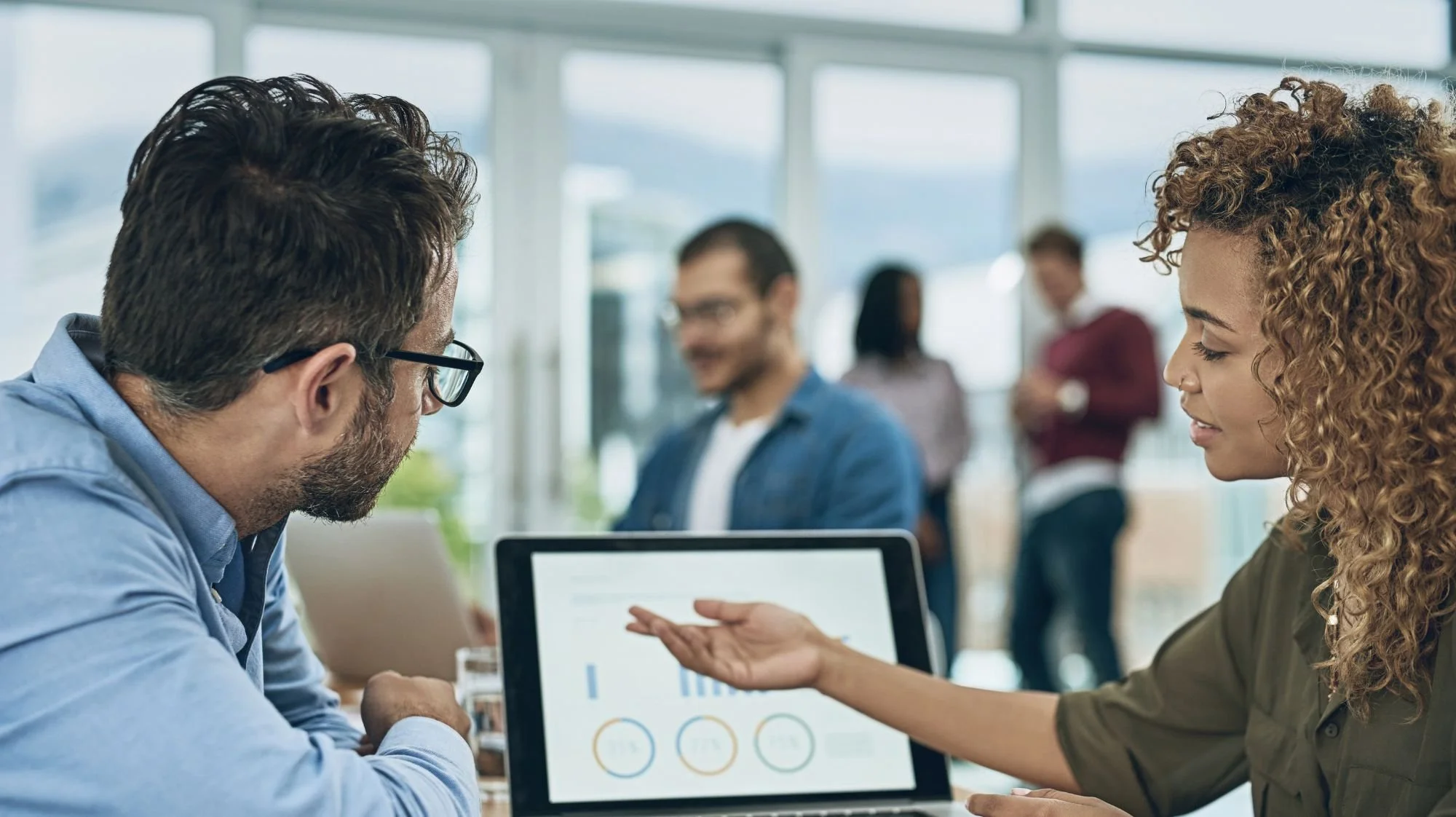 Two people having a business meeting, a man with glasses and a woman with curly hair discussing a tablet with charts, with a blurred office background and other people standing in the background.