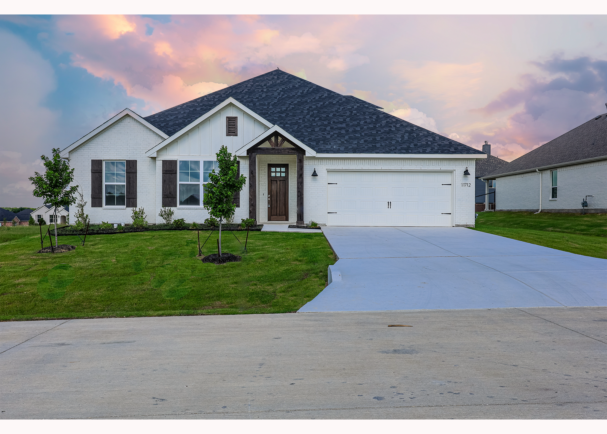 Modern white house with dark roof, large windows, and a newly planted lawn, with a concrete driveway leading to a two-car garage, under a colorful sunset sky