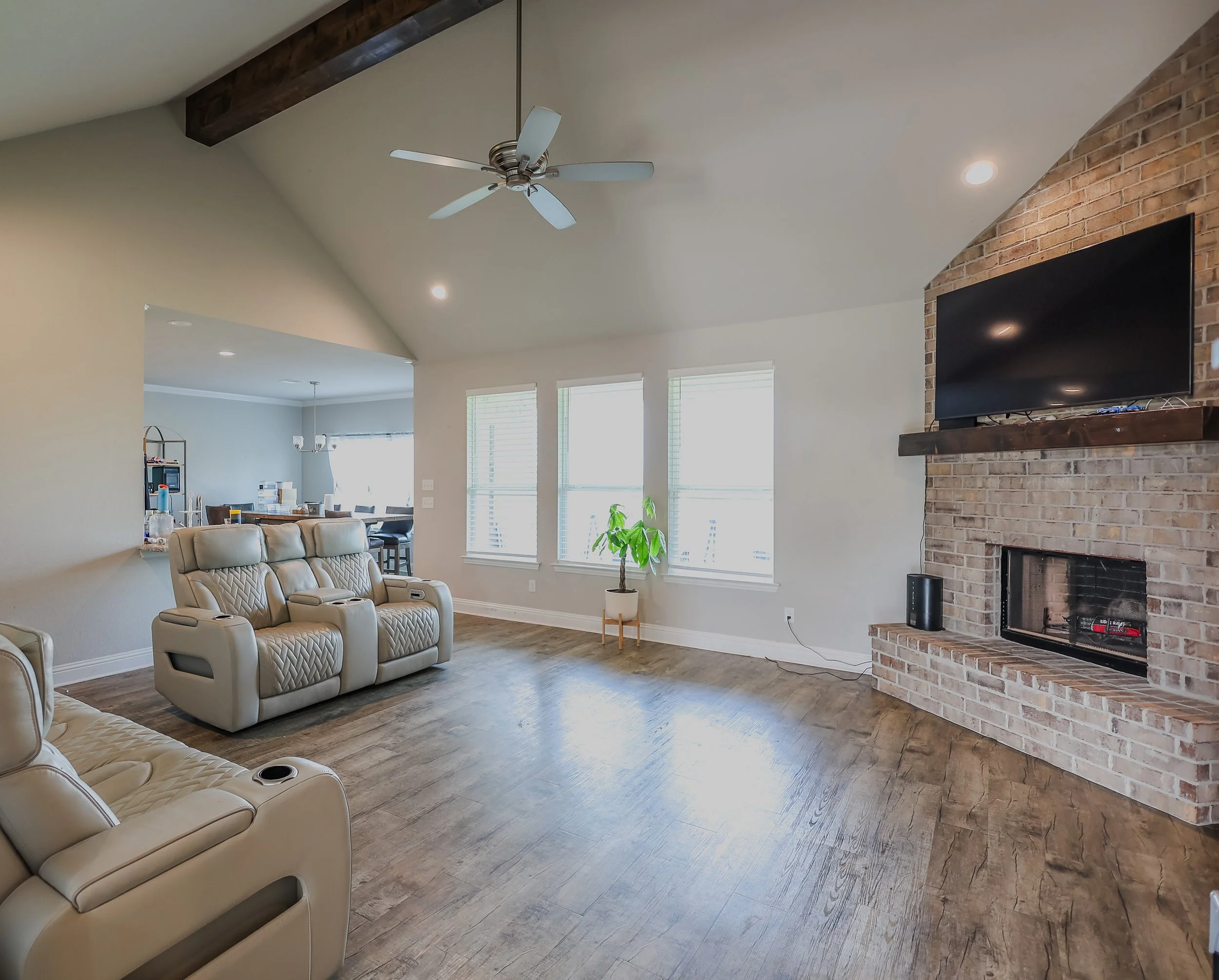 Living room with beige recliner sofas, a brick fireplace with a mounted TV, a green potted plant near windows, and a visible dining area in the background.