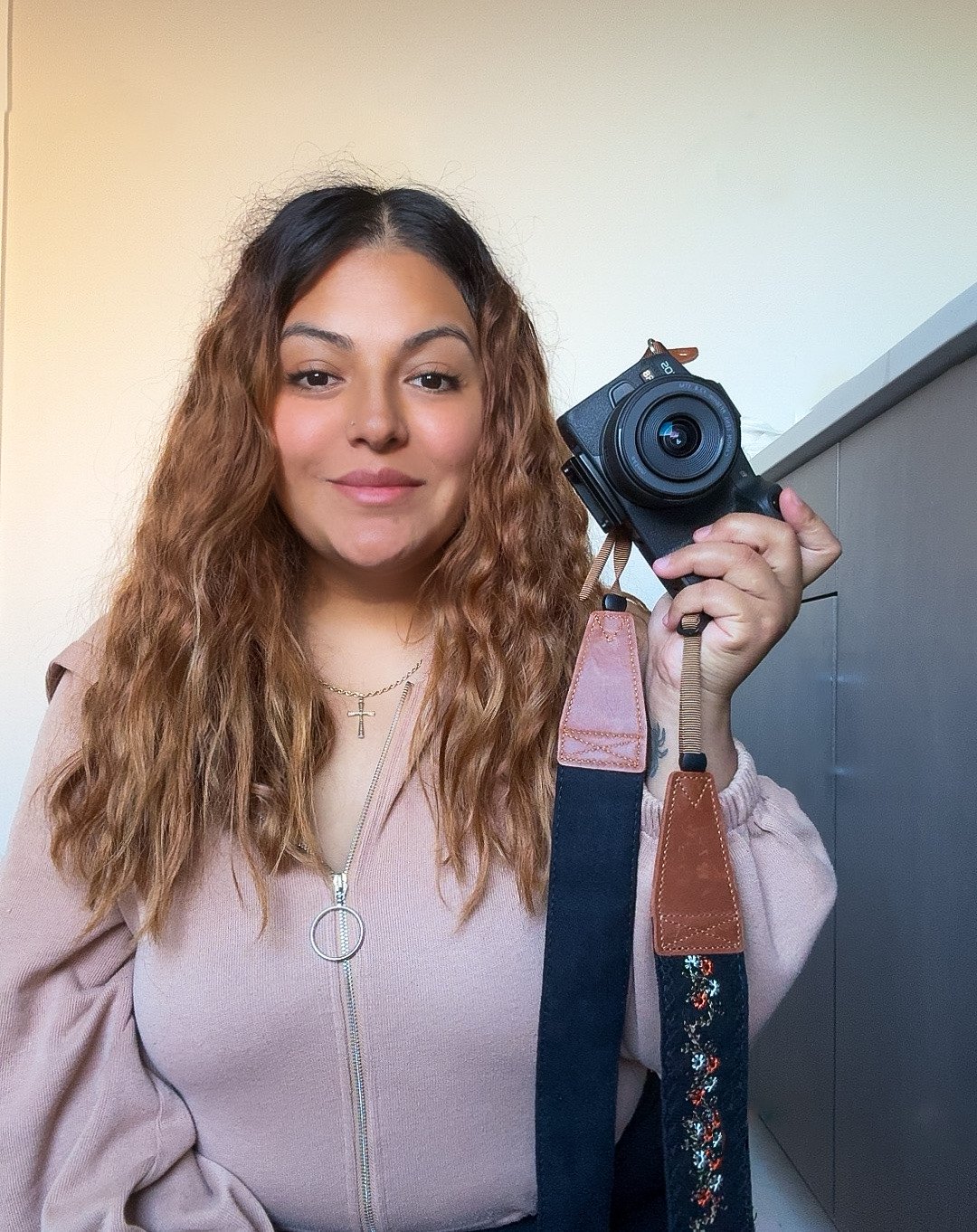 A young woman with wavy light brown hair taking a selfie with a camera in a mirror.