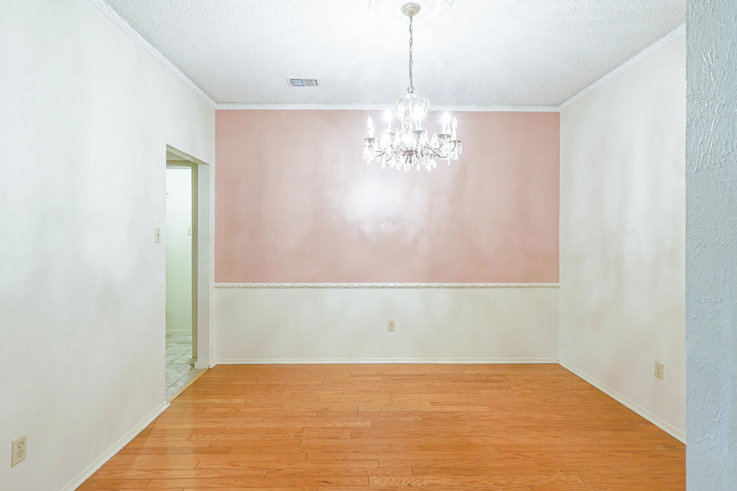 Empty dining room with wood floor, white walls, a pink accent wall, a chandelier hanging from the ceiling, and an open doorway leading to another room.
