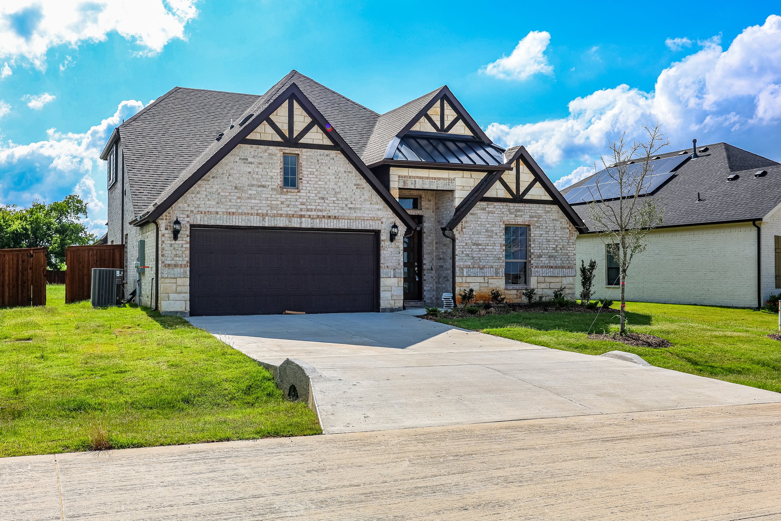 A modern house with brick and stone exterior, black roof, and a two-car garage, situated on a landscaped lawn with a young tree and neighboring houses, under a partly cloudy sky.