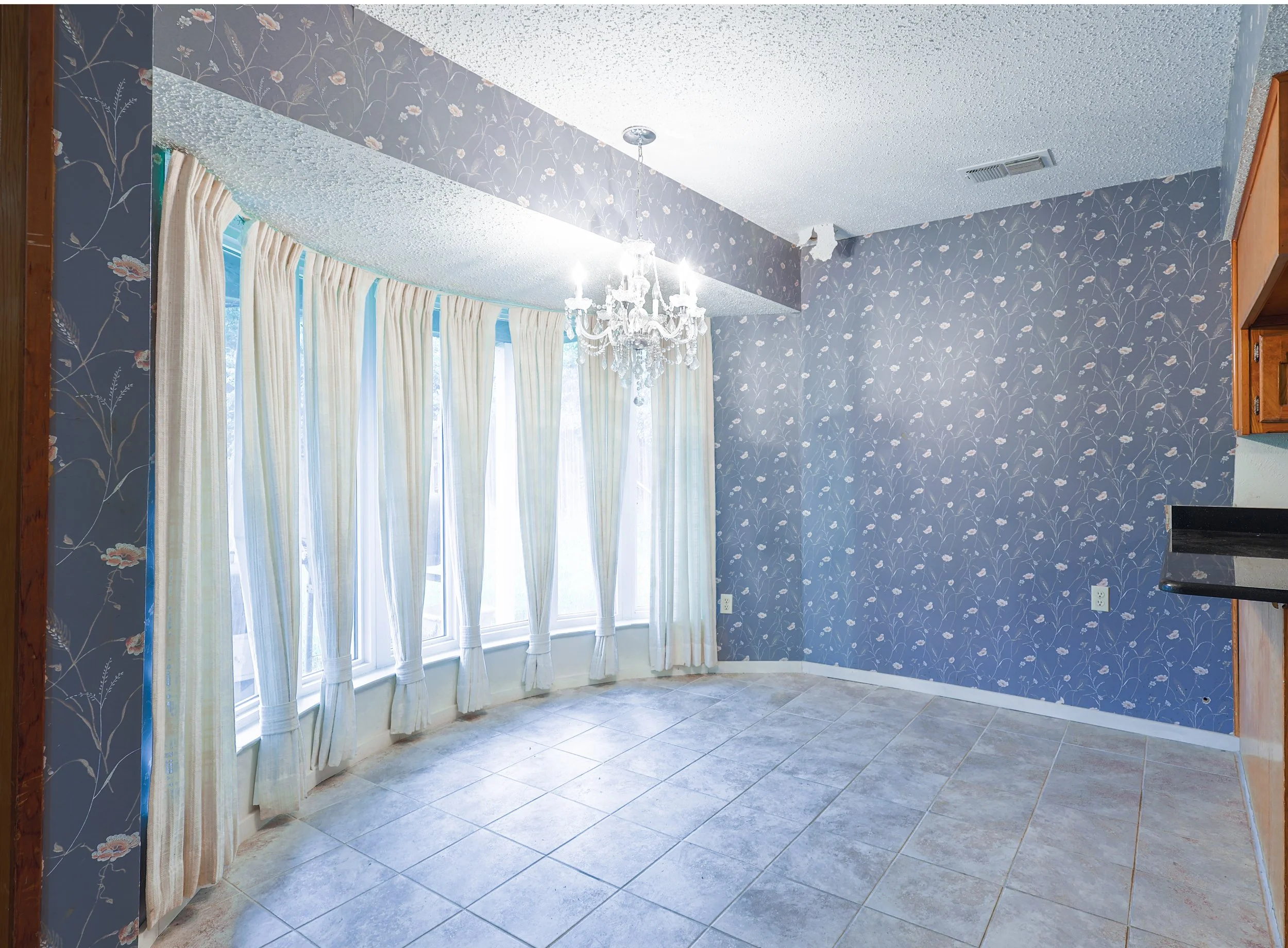Empty room with blue floral wallpaper, beige tiled floor, white curtains on large window, crystal chandelier, and part of a kitchen counter on the right.