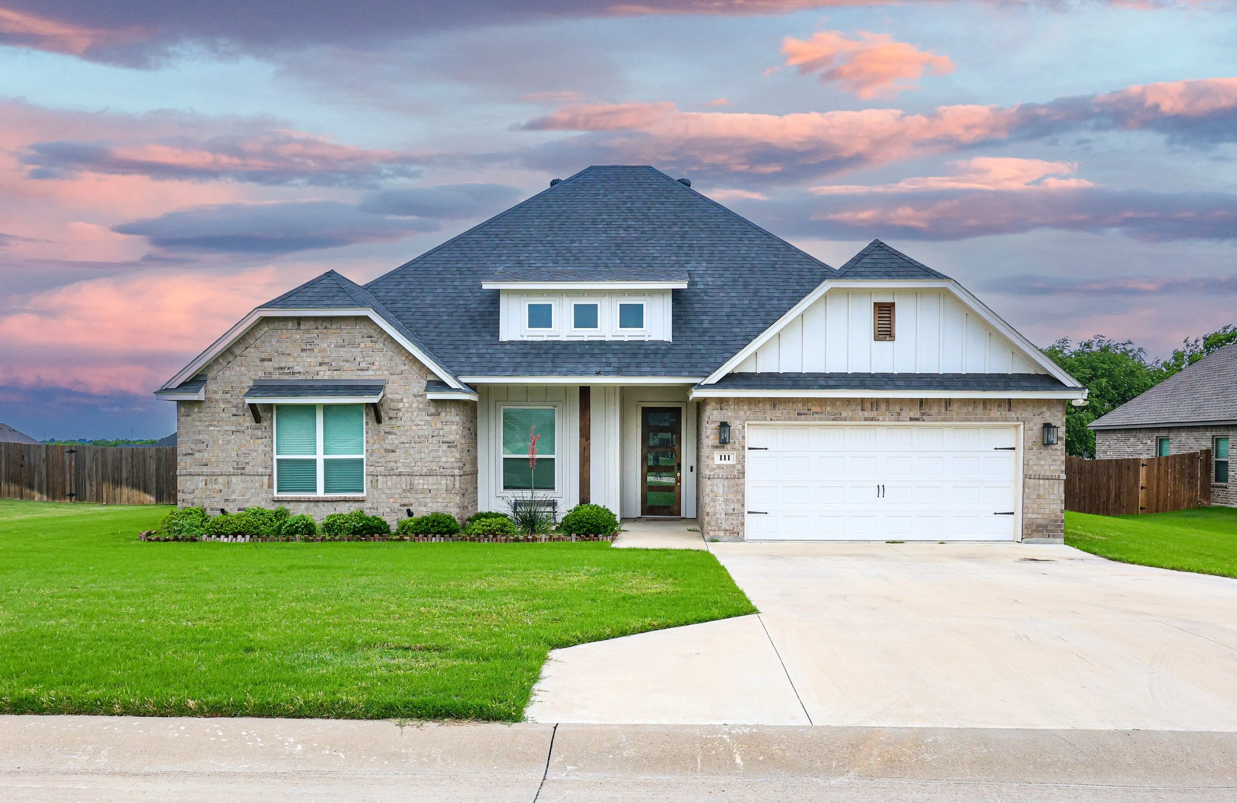 Front view of a modern suburban house with brick and white siding exterior, a two-car garage, and a well-maintained green lawn under a sunset sky.