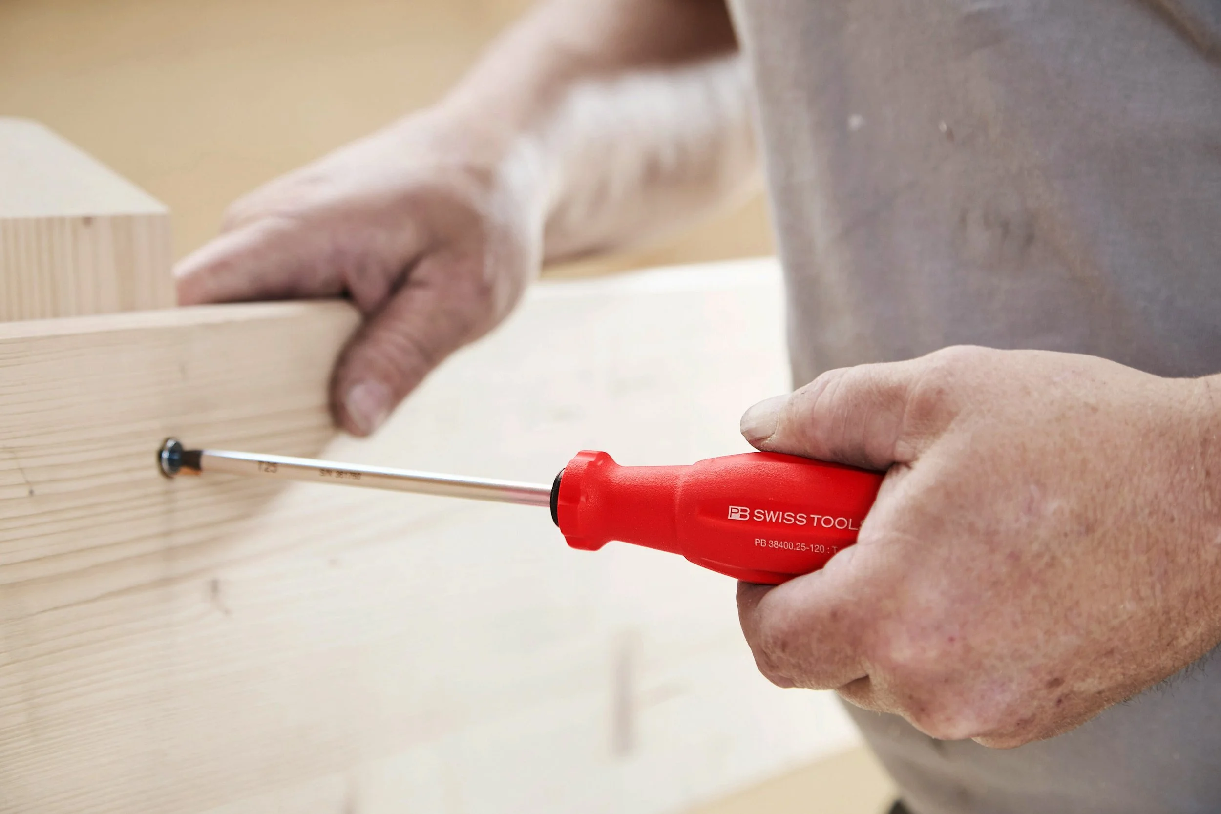 A person using a red screwdriver to assemble light-colored wooden furniture in a workshop.