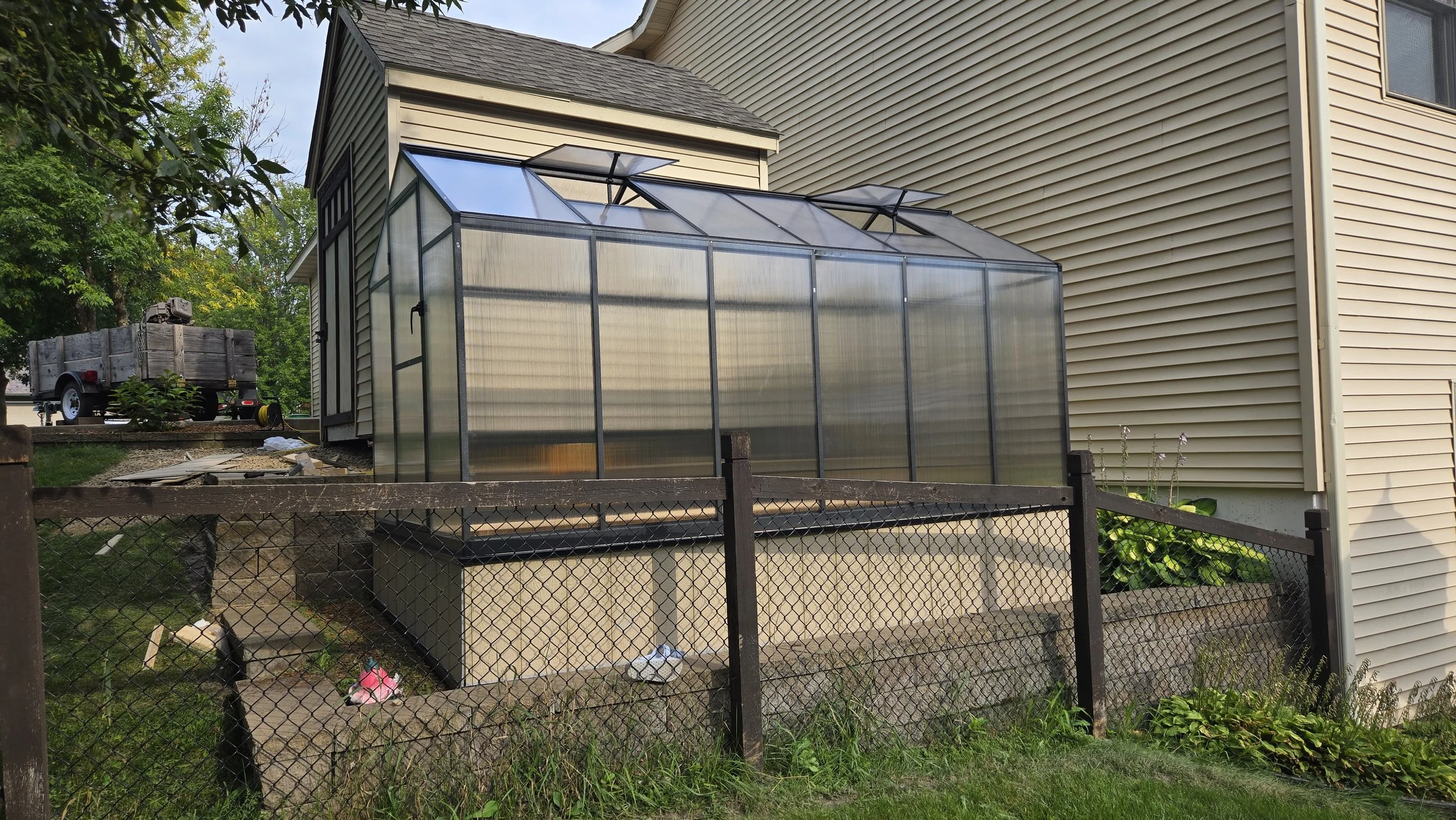 A greenhouse attached to the side of a house with beige siding, surrounded by a black chain-link fence, with a small garden bed and plants nearby.