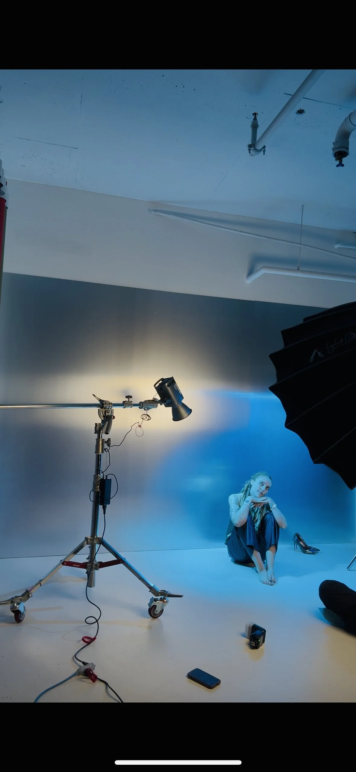 A woman sitting on the floor in a photography studio with a blue background, surrounded by studio lighting equipment, a remote control, and a shoe placed on the floor nearby.
