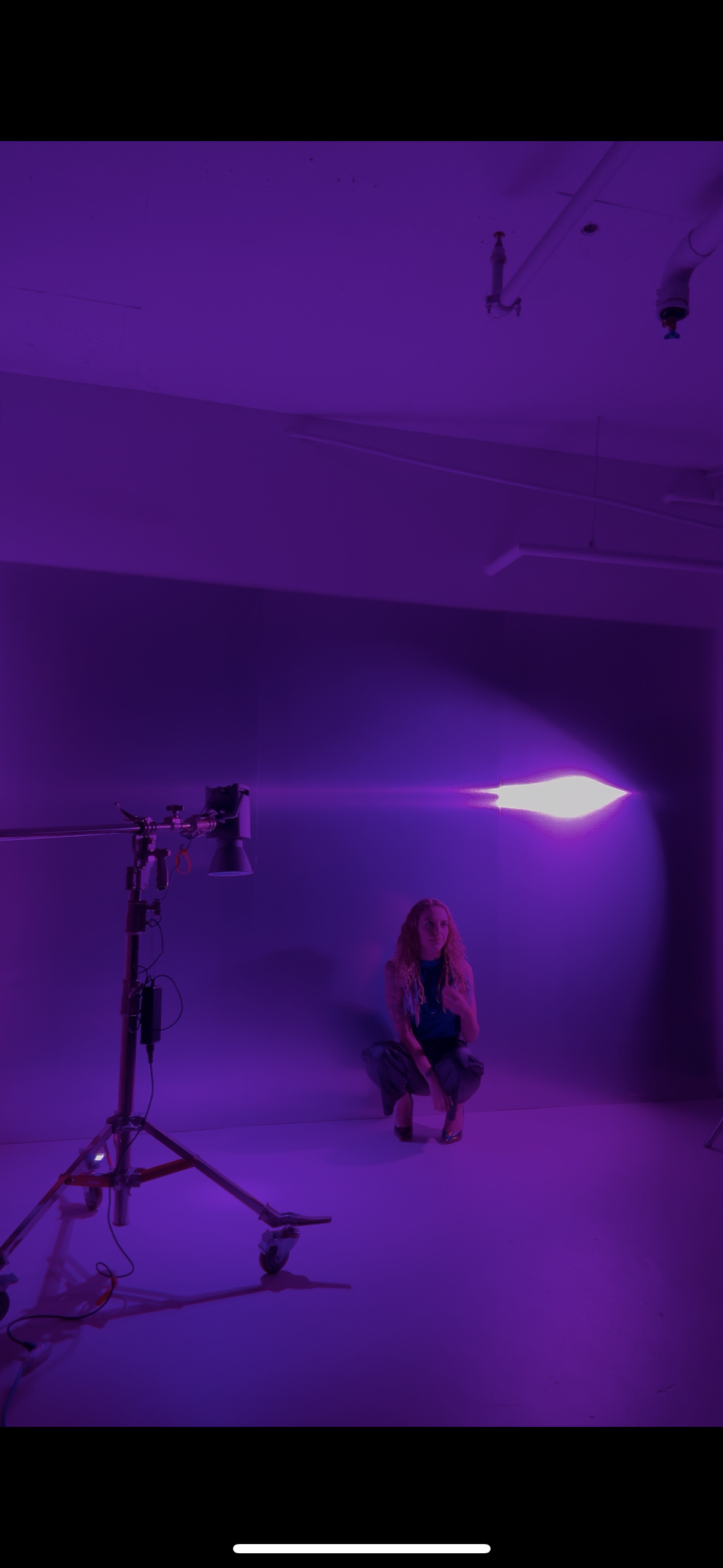 A woman squatting in a dark room with purple lighting, standing next to a large studio light mounted on a stand.