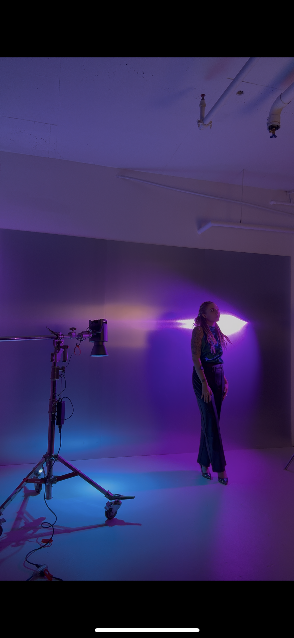 A woman with pink and purple hair standing in a studio with violet and purple lighting, next to equipment on a wheeled stand and a violet-colored spotlight hitting the wall.