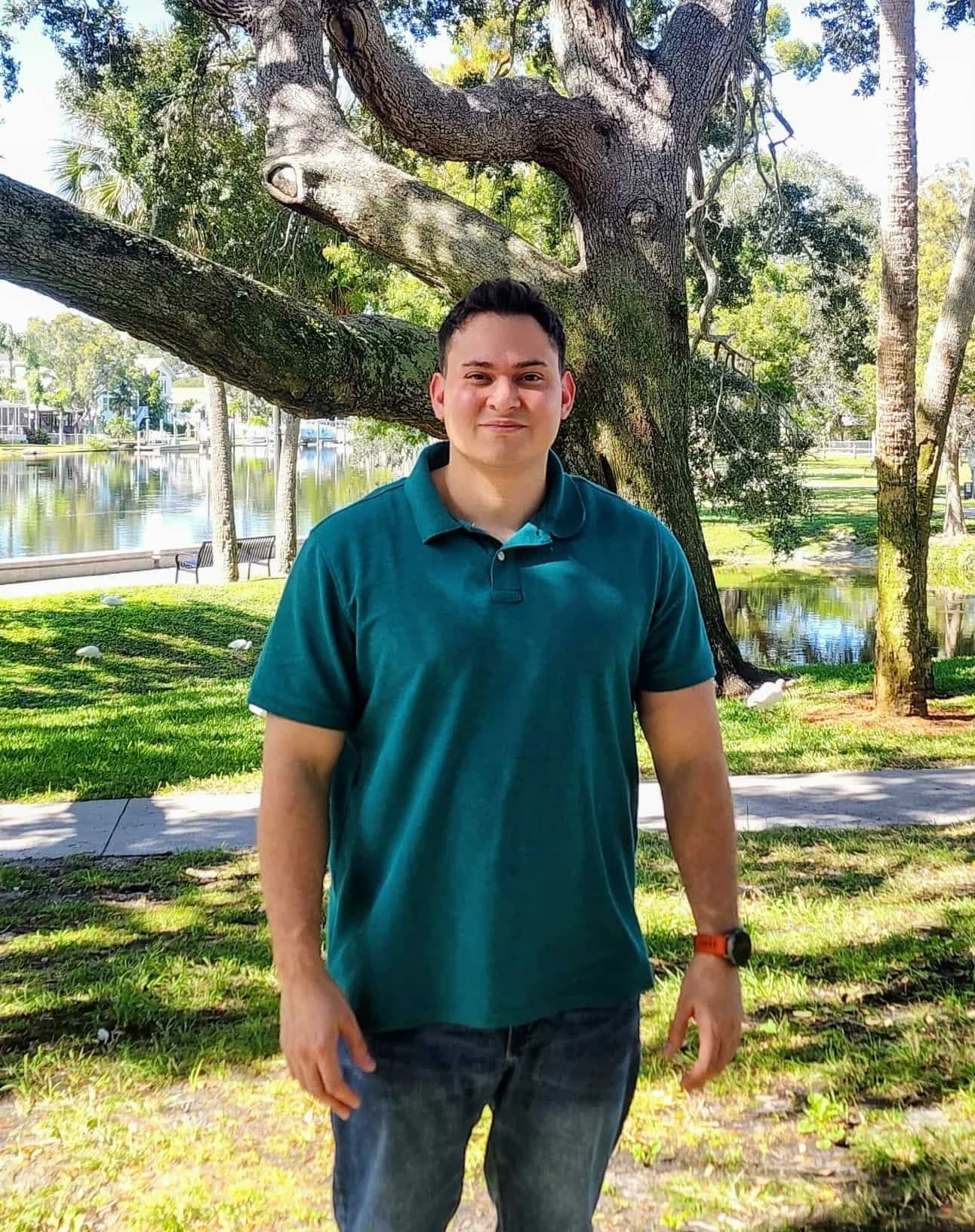 A young man standing outdoors by a large tree near a body of water, wearing a teal polo shirt and jeans.