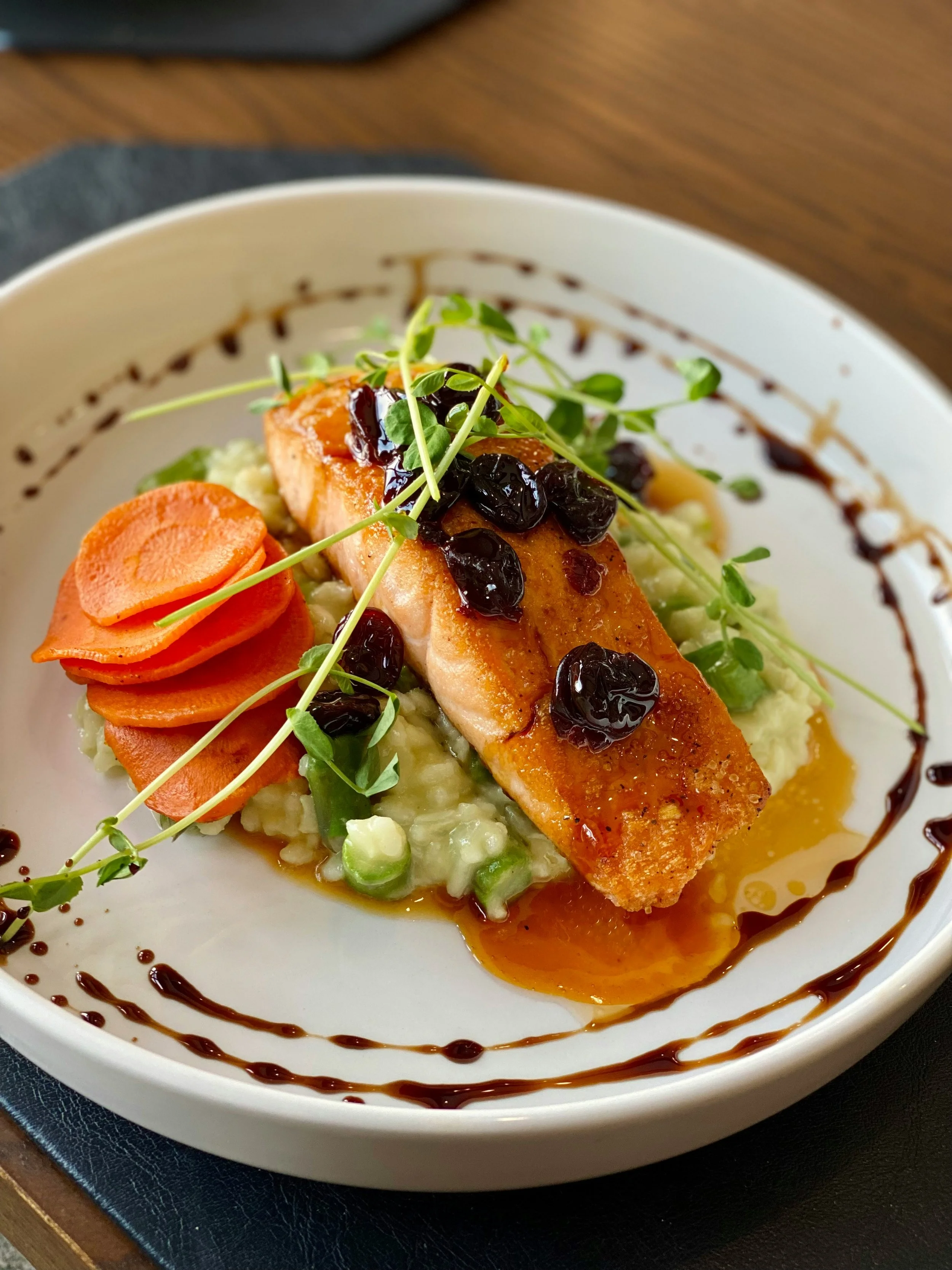 Seared salmon fillet garnished with microgreens and blackberries, served over mashed potatoes, with sliced carrots and a drizzle of balsamic glaze around the plate.