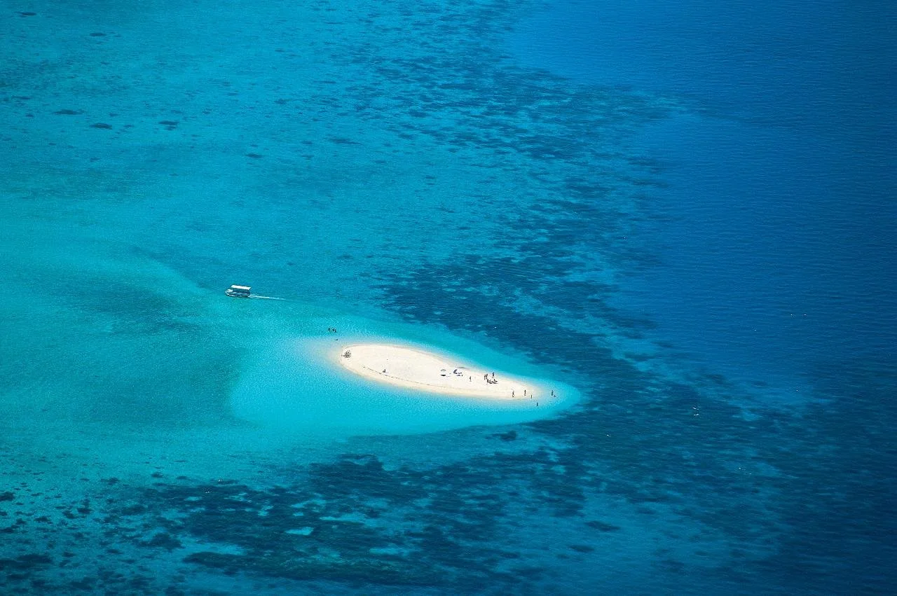 An aerial view of a small, sandy island with people on it, surrounded by clear turquoise water. A boat is near the island, creating a wake.
