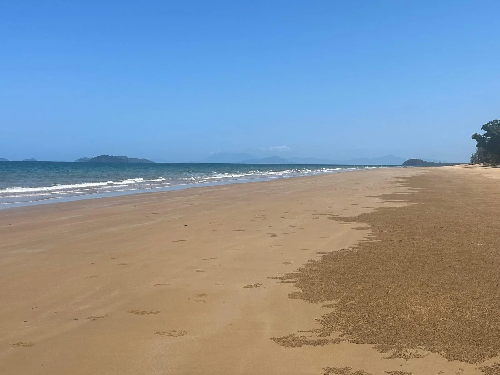 Wide view of a sandy beach with gentle waves and distant islands under a clear blue sky.