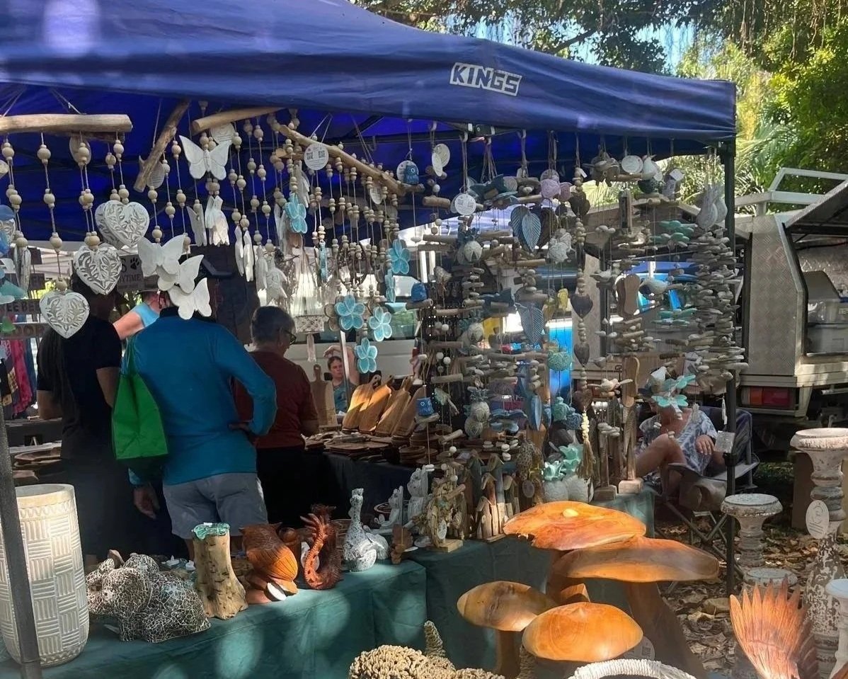 Outdoor craft market stall with wooden and ceramic decor, including animals, mushrooms, butterflies, and hanging ornaments, under a blue canopy, with visitors browsing.