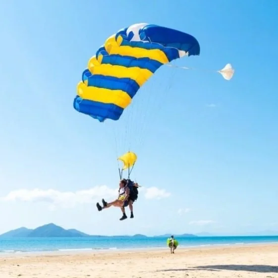 A person parasailing with a yellow and blue parachute on a sandy beach, with two other people walking in the distance and an island in the background under a clear blue sky.