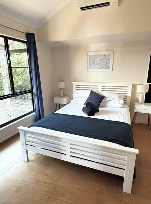 Bedroom with white bed frame, black blanket, black pillow, and rolled black towels, two nightstands with lamps, window with blue curtain, and beach-themed artwork on wall.