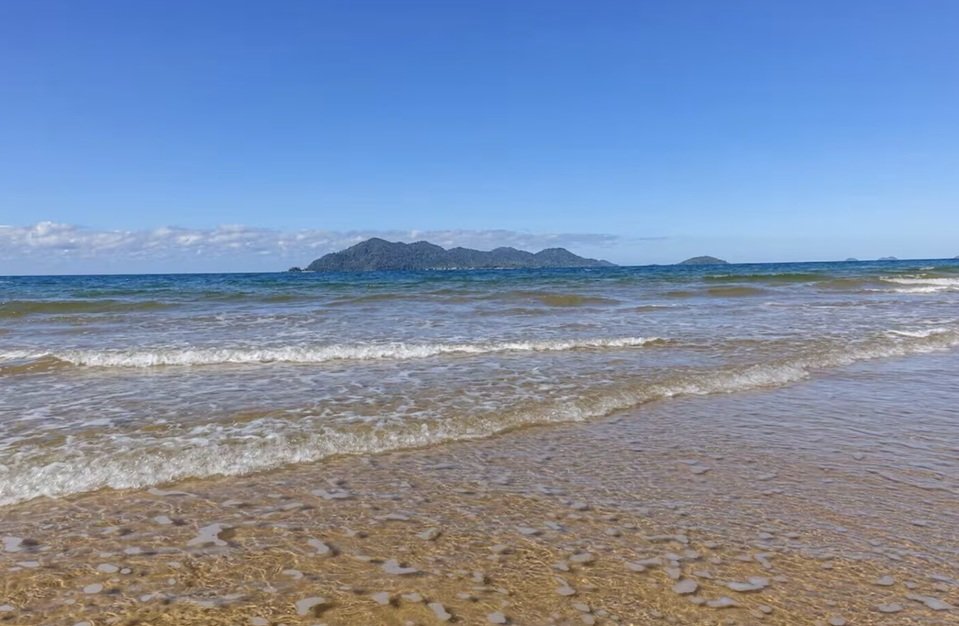 A beach scene with gentle waves on a sandy shore, and an island with hills in the distance under a clear blue sky.